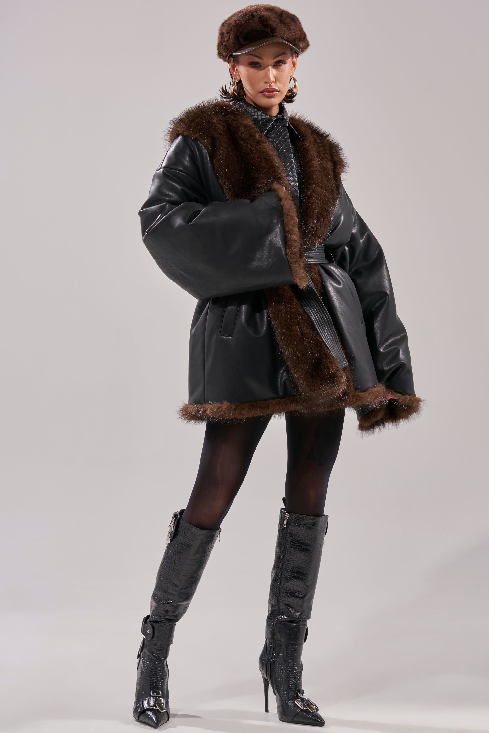 A woman models the CAN I KICK IT REVERSIBLE PUFFER—a black faux leather coat with brown fur trim—paired with patterned tights, textured knee-high heeled boots, and a matching brown fur beret against a neutral background.