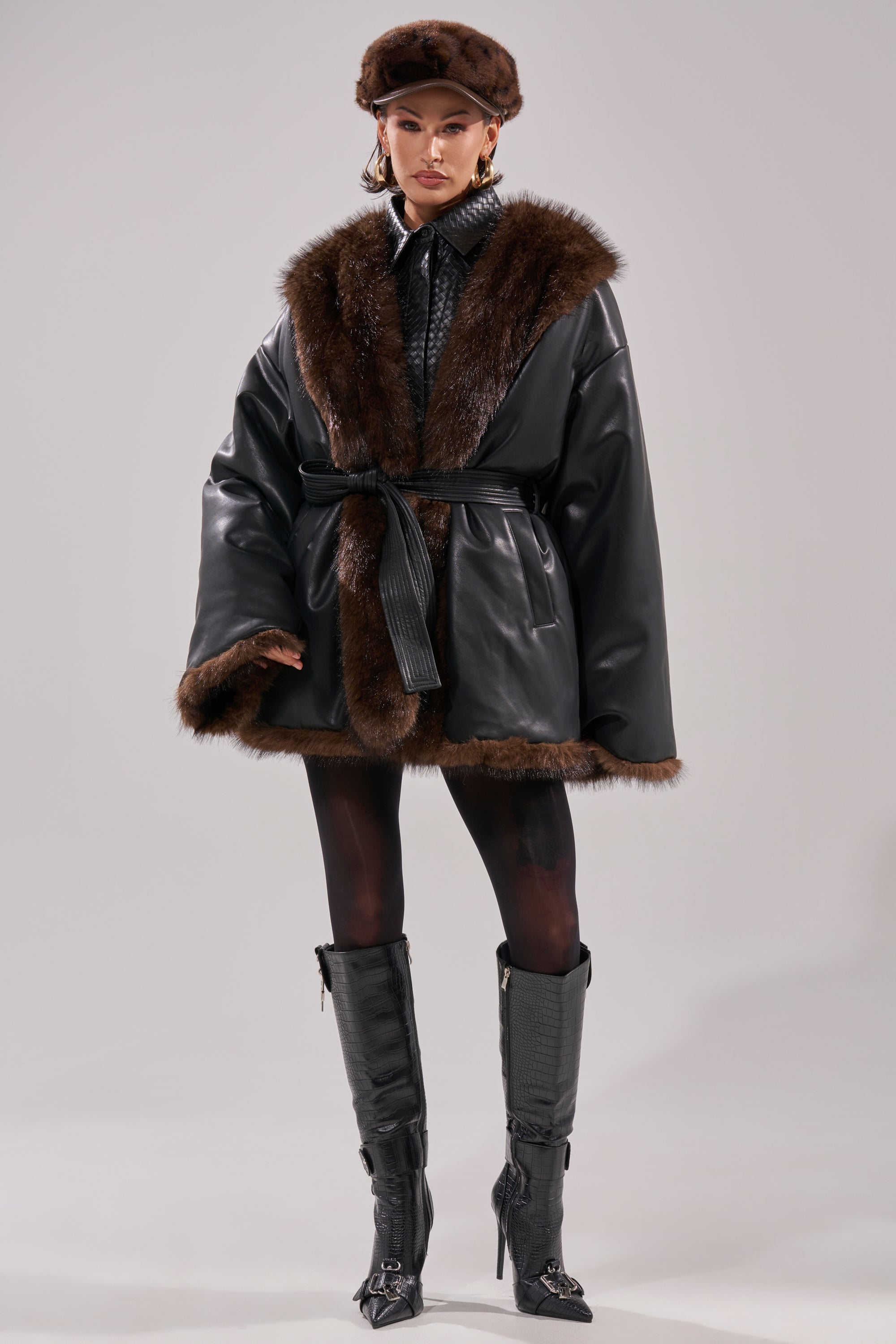A woman stands facing forward in the CAN I KICK IT REVERSIBLE PUFFER, styled with a matching fur hat, black patterned tights, knee-high black boots, and gold hoop earrings against a plain background.