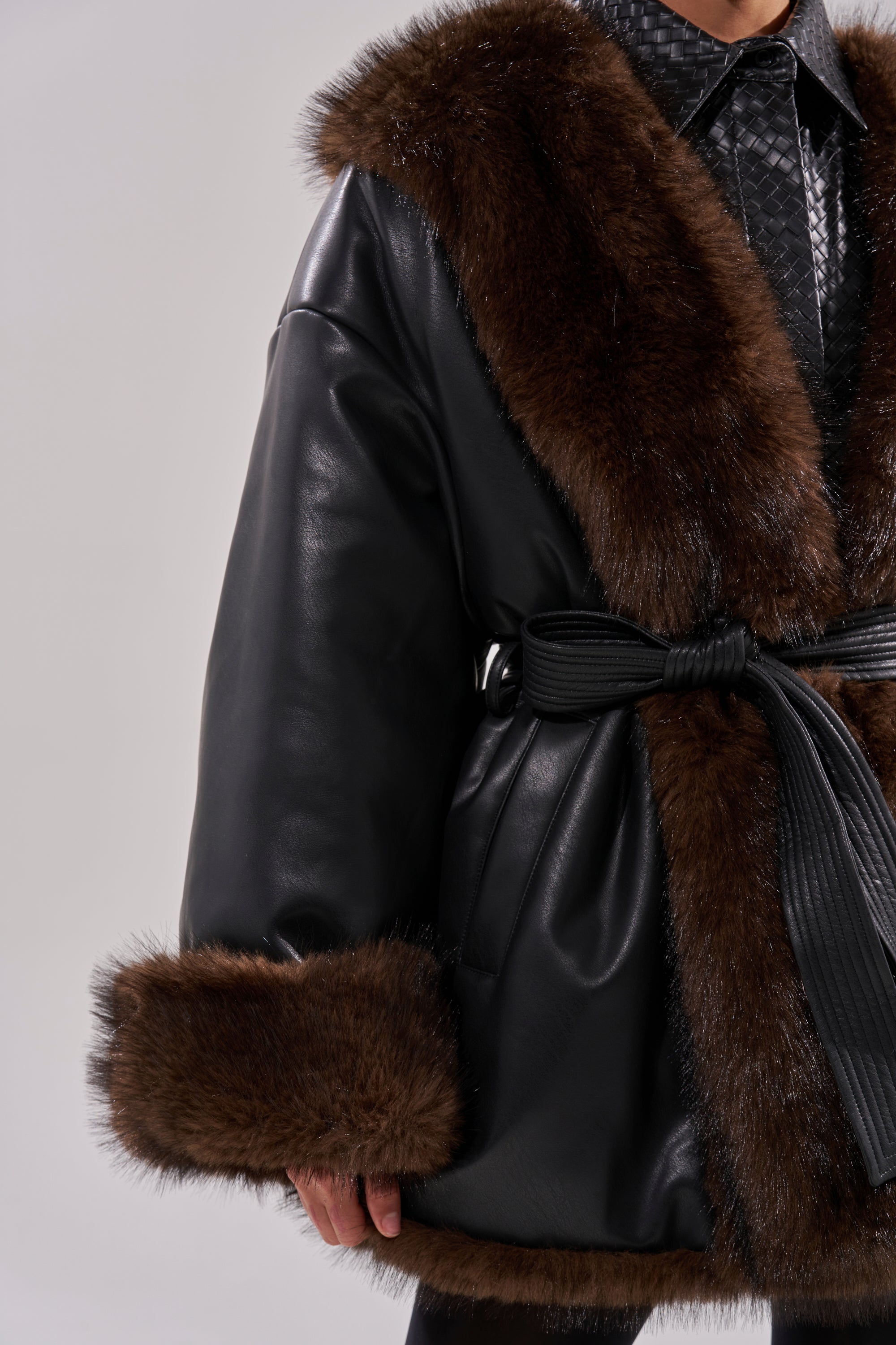 A person wears the CAN I KICK IT REVERSIBLE PUFFER—a black faux leather coat with wide dark brown faux fur trim on the collar and cuffs, cinched at the waist with a textured black belt. The background is neutral and uncluttered.