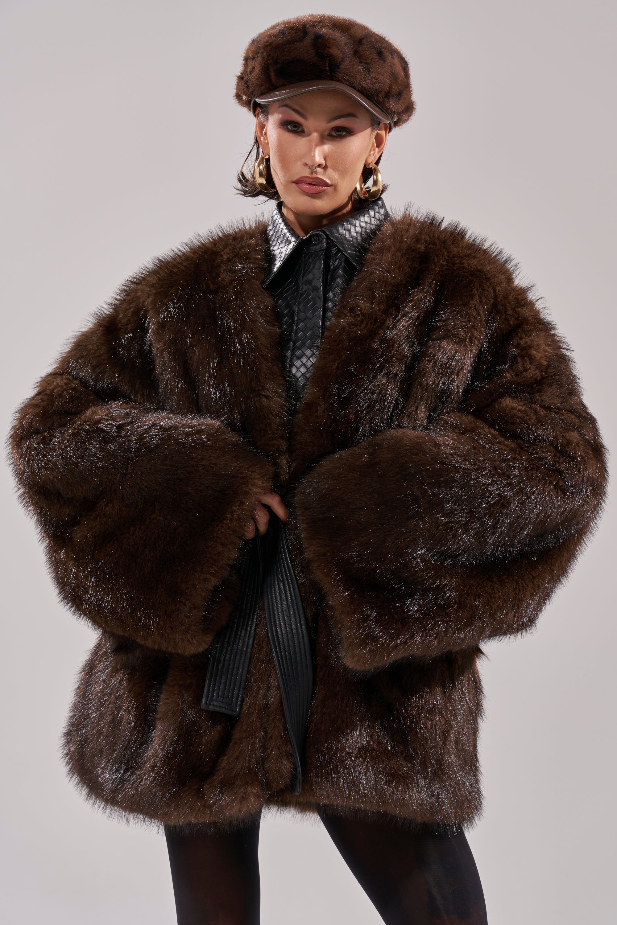 A person models the CAN I KICK IT REVERSIBLE PUFFER, styled with a matching fur hat, hoop earrings, and collared shirt, hands near the opening, highlighting the plush appeal of this dark brown faux fur coat against a plain background.