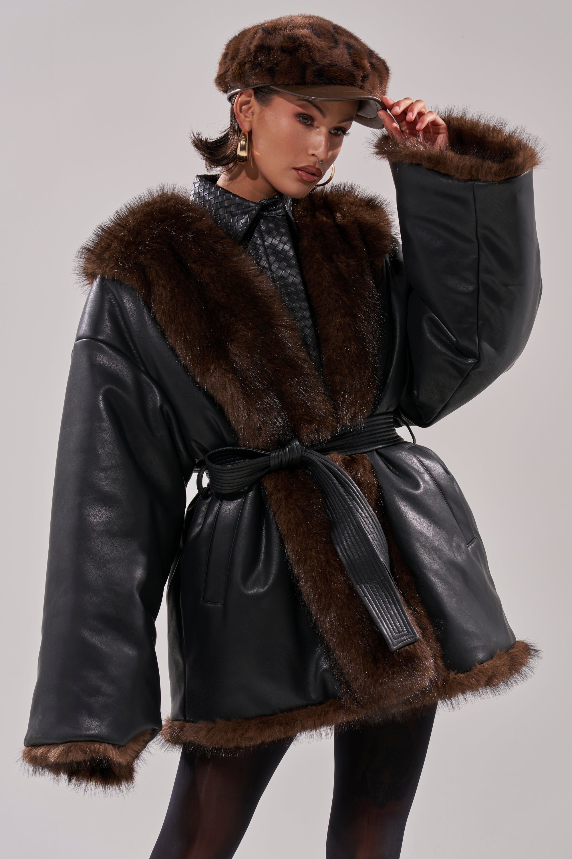 A woman models the CAN I KICK IT REVERSIBLE PUFFER, a black faux leather coat with wide sleeves, faux fur trim, and a belted waist, styled with a matching fur hat and gold hoops against a plain background.