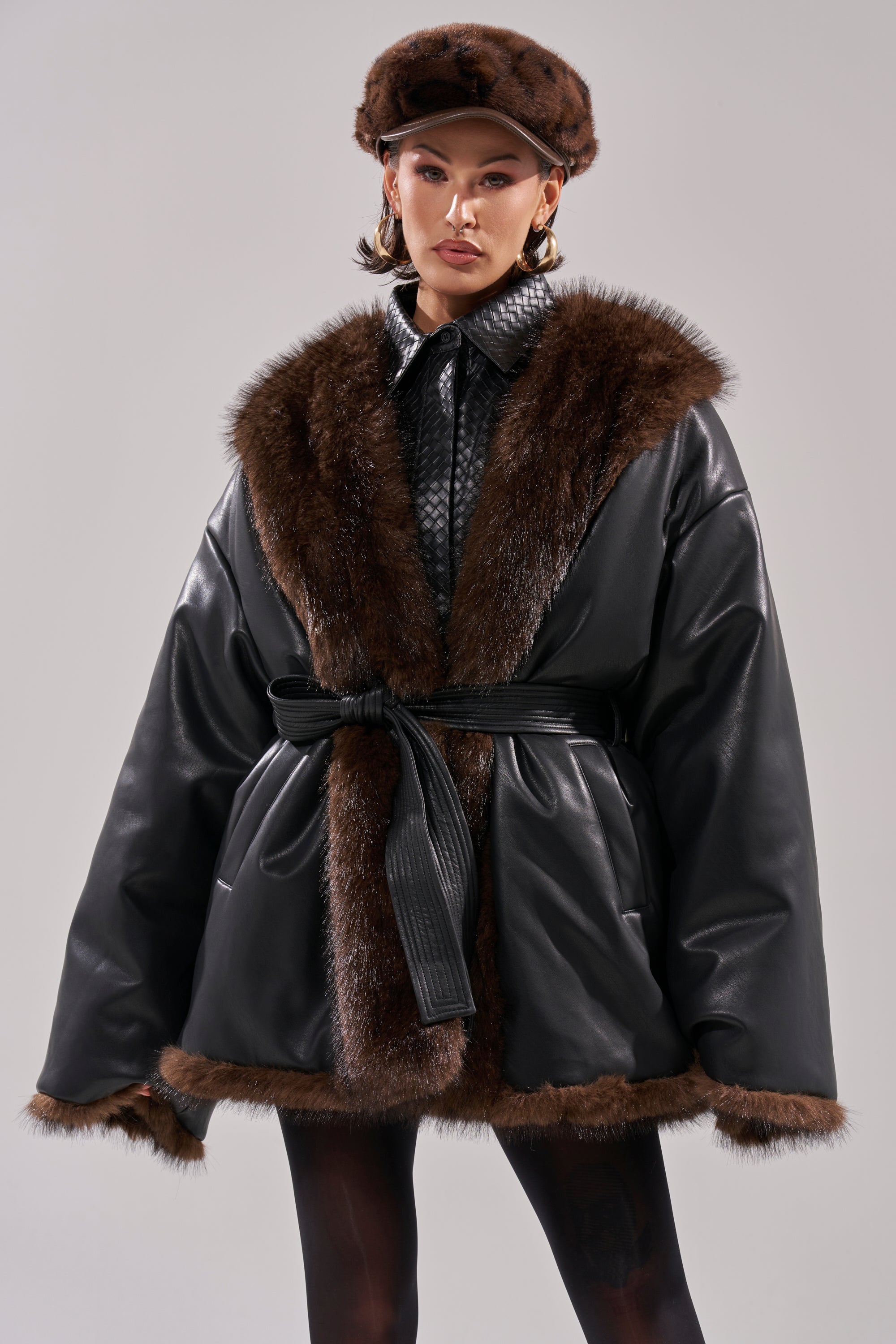A person wearing the CAN I KICK IT REVERSIBLE PUFFER, a brown fur hat, large hoop earrings, and patterned black tights stands facing forward against a plain light background.