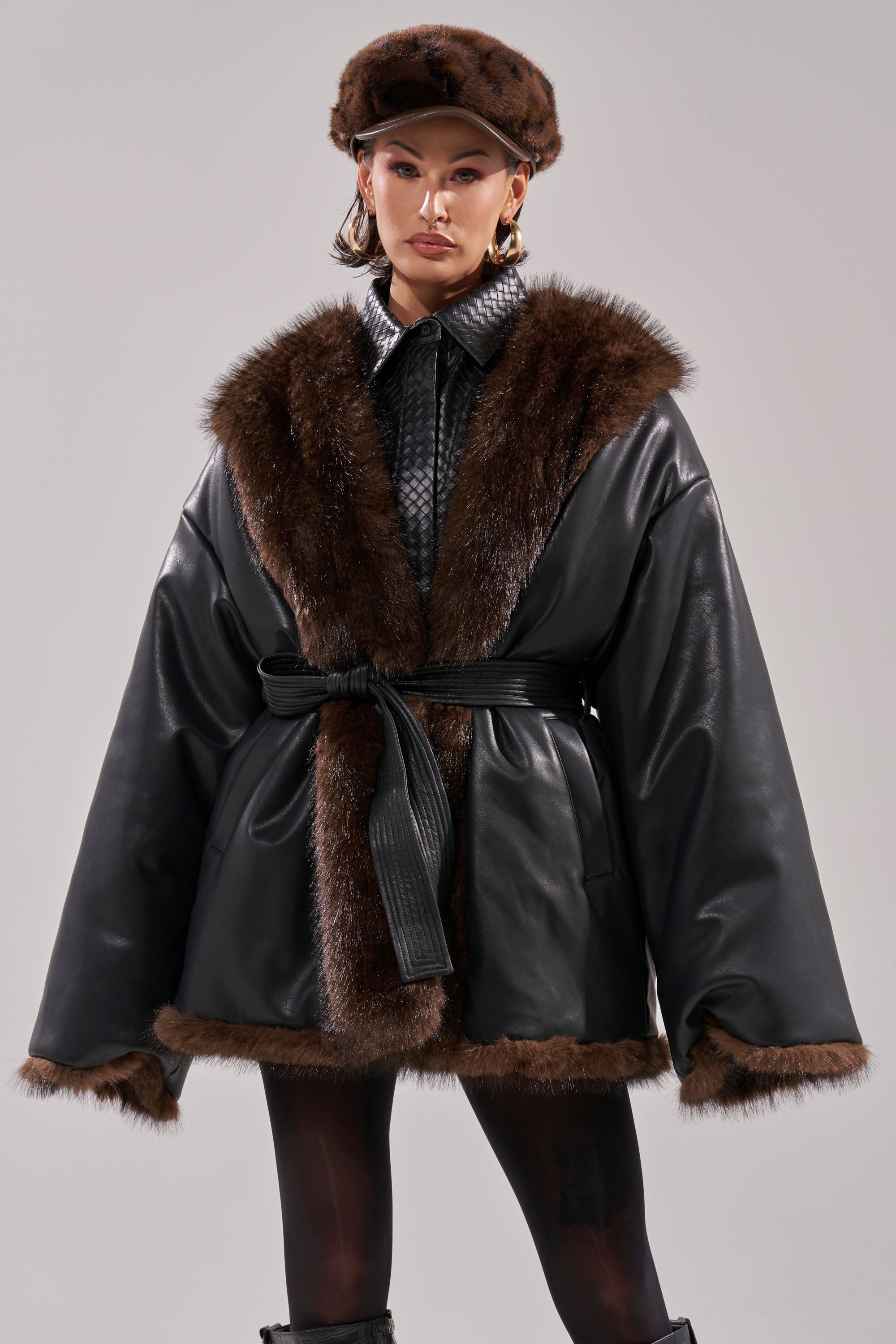 A woman stands against a plain background wearing the CAN I KICK IT REVERSIBLE PUFFER with brown faux fur trim, a matching fur hat, hoop earrings, and a textured black shirt under the belted coat.