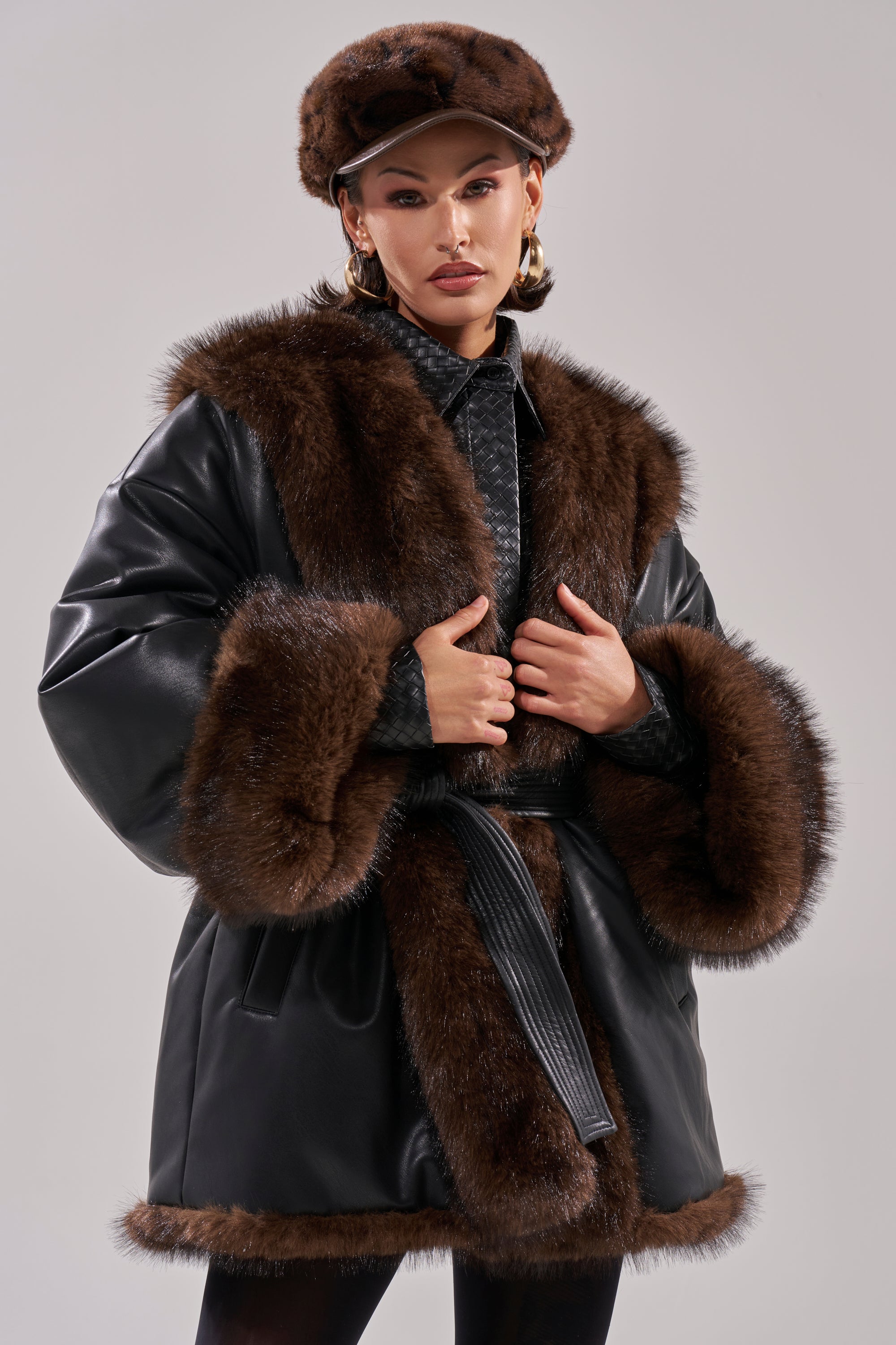 Woman in the CAN I KICK IT REVERSIBLE PUFFER, featuring wide dark brown faux fur trim and a matching hat, gold hoop earrings, and dark lipstick, posing against a neutral gray background.