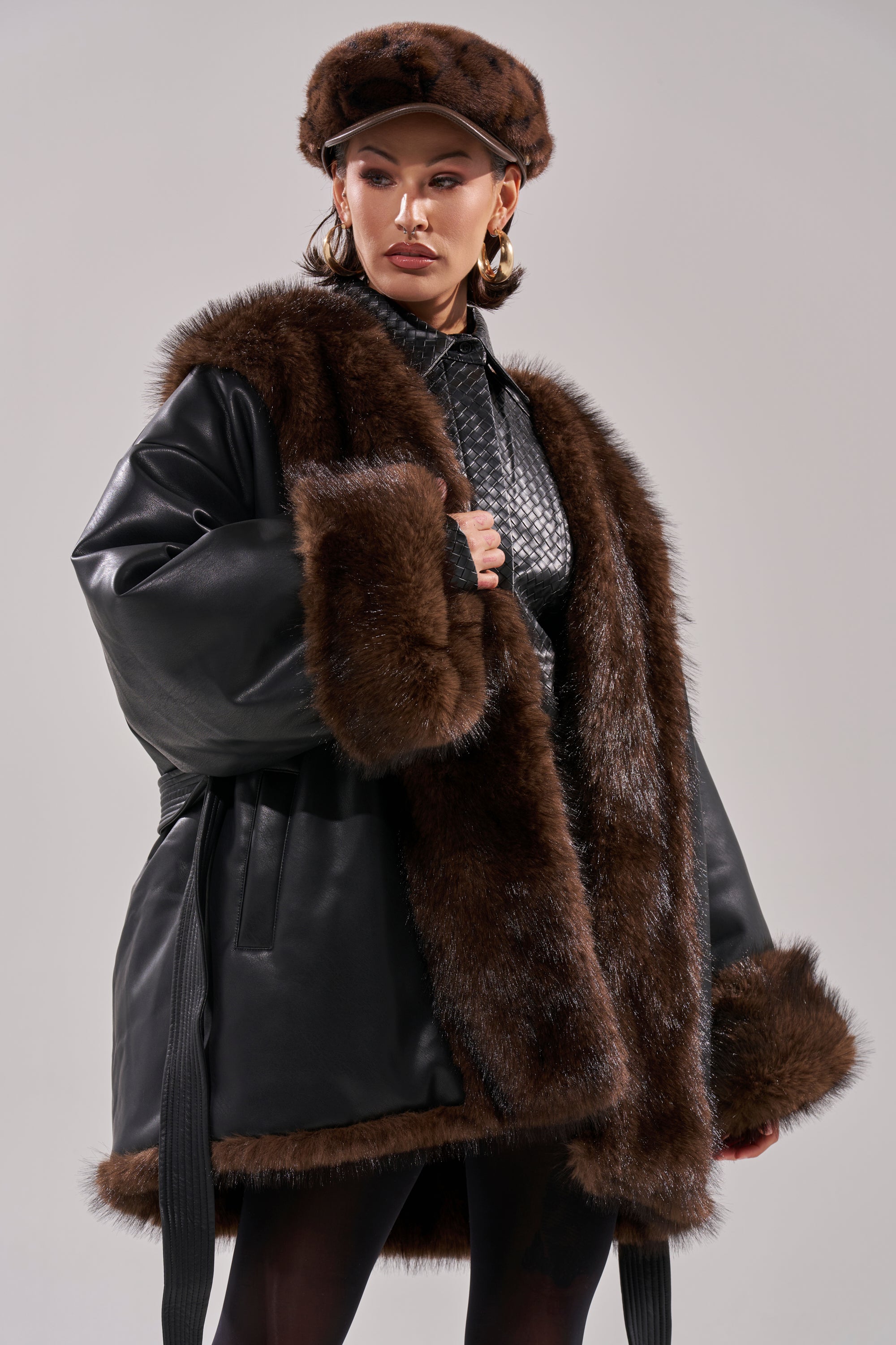 A person wearing the CAN I KICK IT REVERSIBLE PUFFER, styled with a matching fur hat, black patterned shirt, and dark stockings, poses confidently against a plain light gray background.