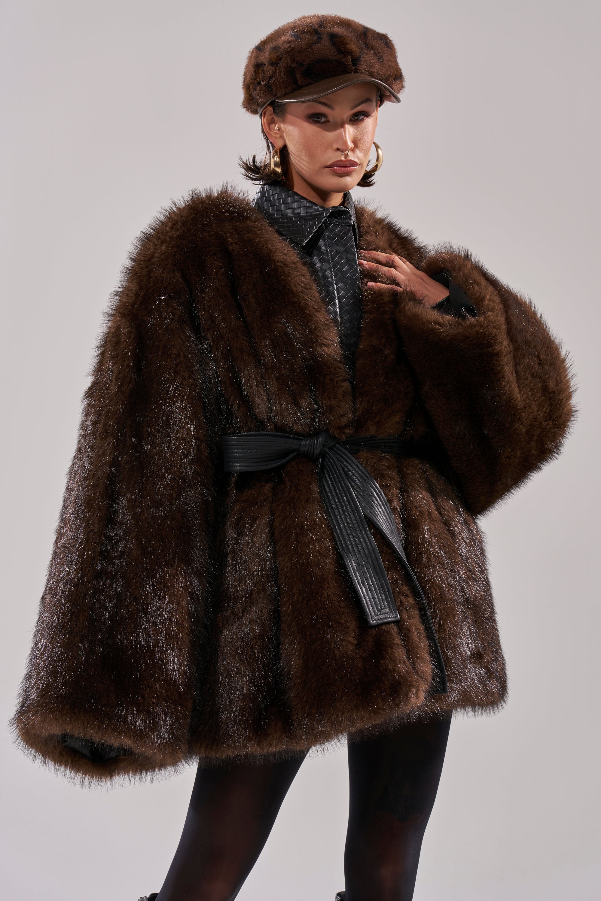 Wearing the CAN I KICK IT REVERSIBLE PUFFER, a person pairs it with a black belt, matching brown fur hat, hoop earrings, and a black collared shirt while posing confidently against a plain background.