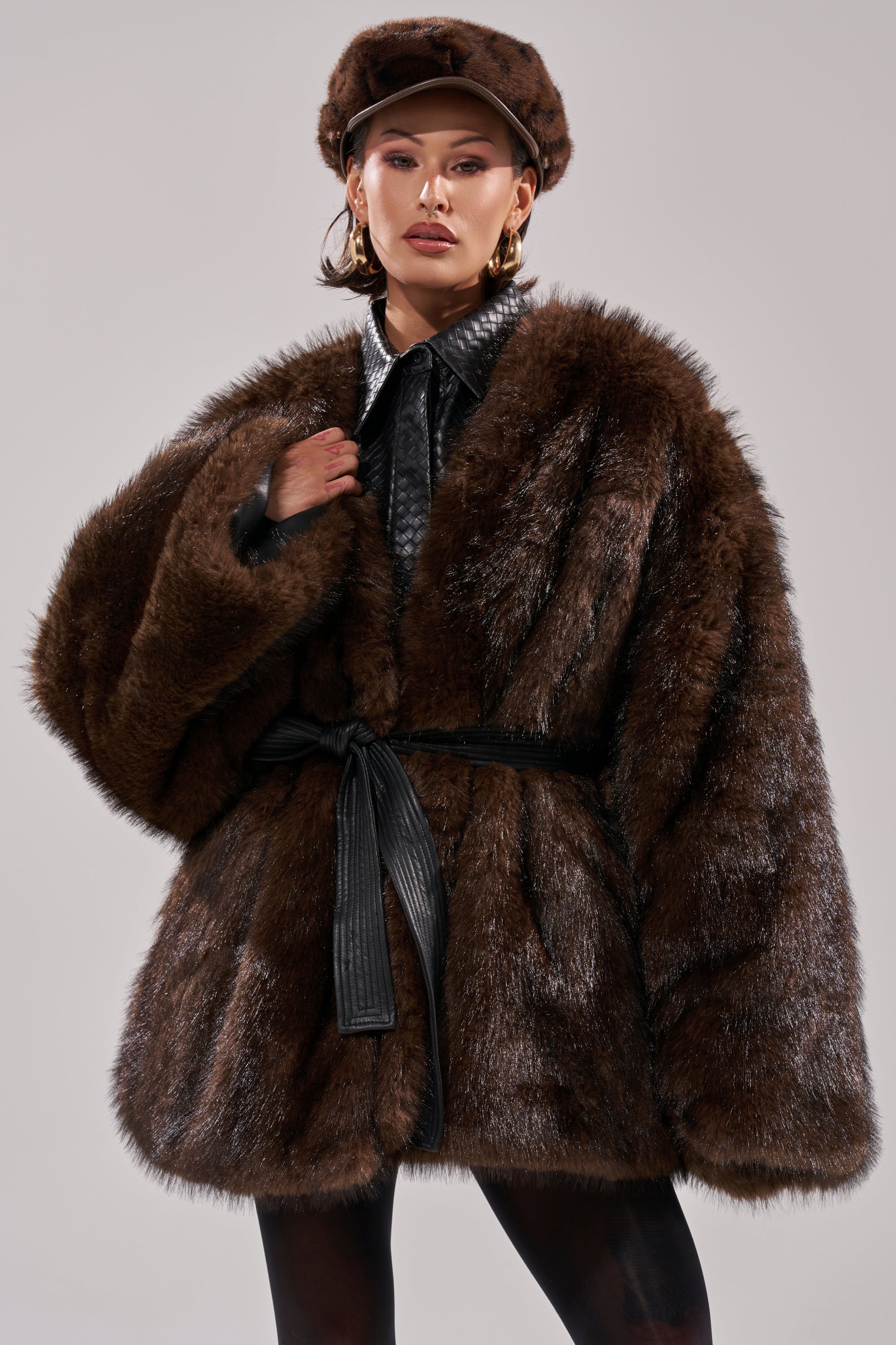 A woman models the CAN I KICK IT REVERSIBLE PUFFER in dark brown, styled with a black belt, matching fur hat, hoop earrings, and collared shirt against a neutral background for a chic winter look.