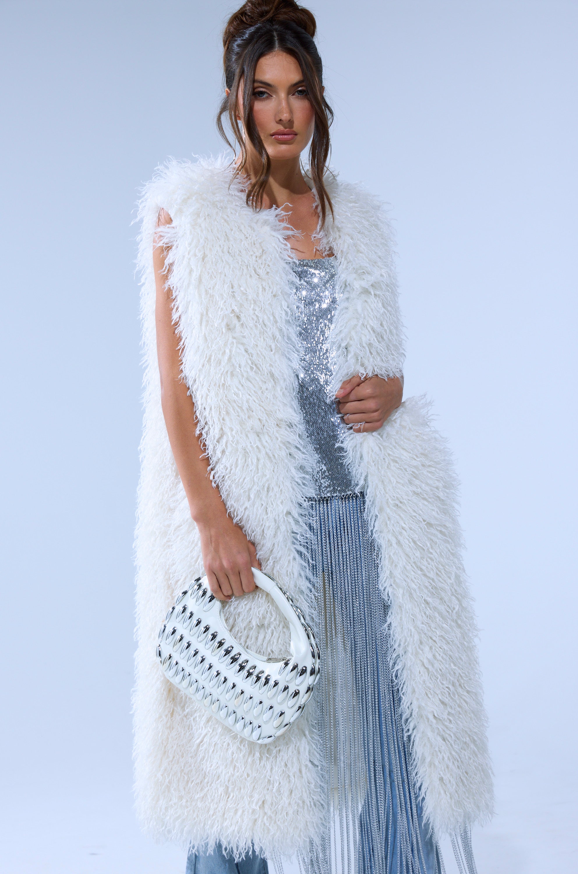 A woman models the ANOTHER LANE MOHAIR VEST, a longline white faux fur piece, over a shimmering silver top and pleated skirt, holding a white handbag with black accents against a plain light background.