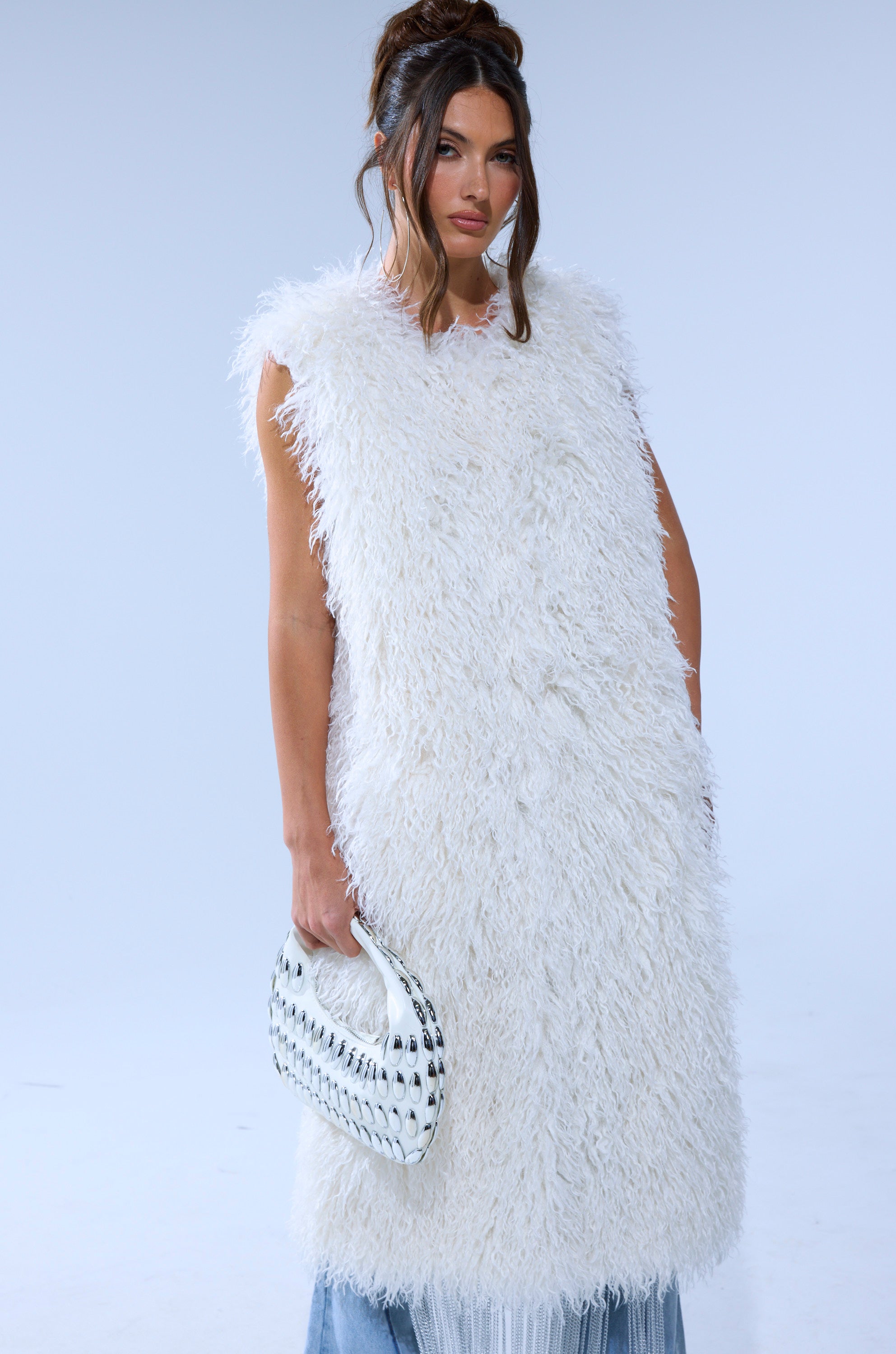 A woman with long brown hair in an updo wears the sleeveless white ANOTHER LANE MOHAIR VEST and holds a small white handbag, standing against a plain light blue background.