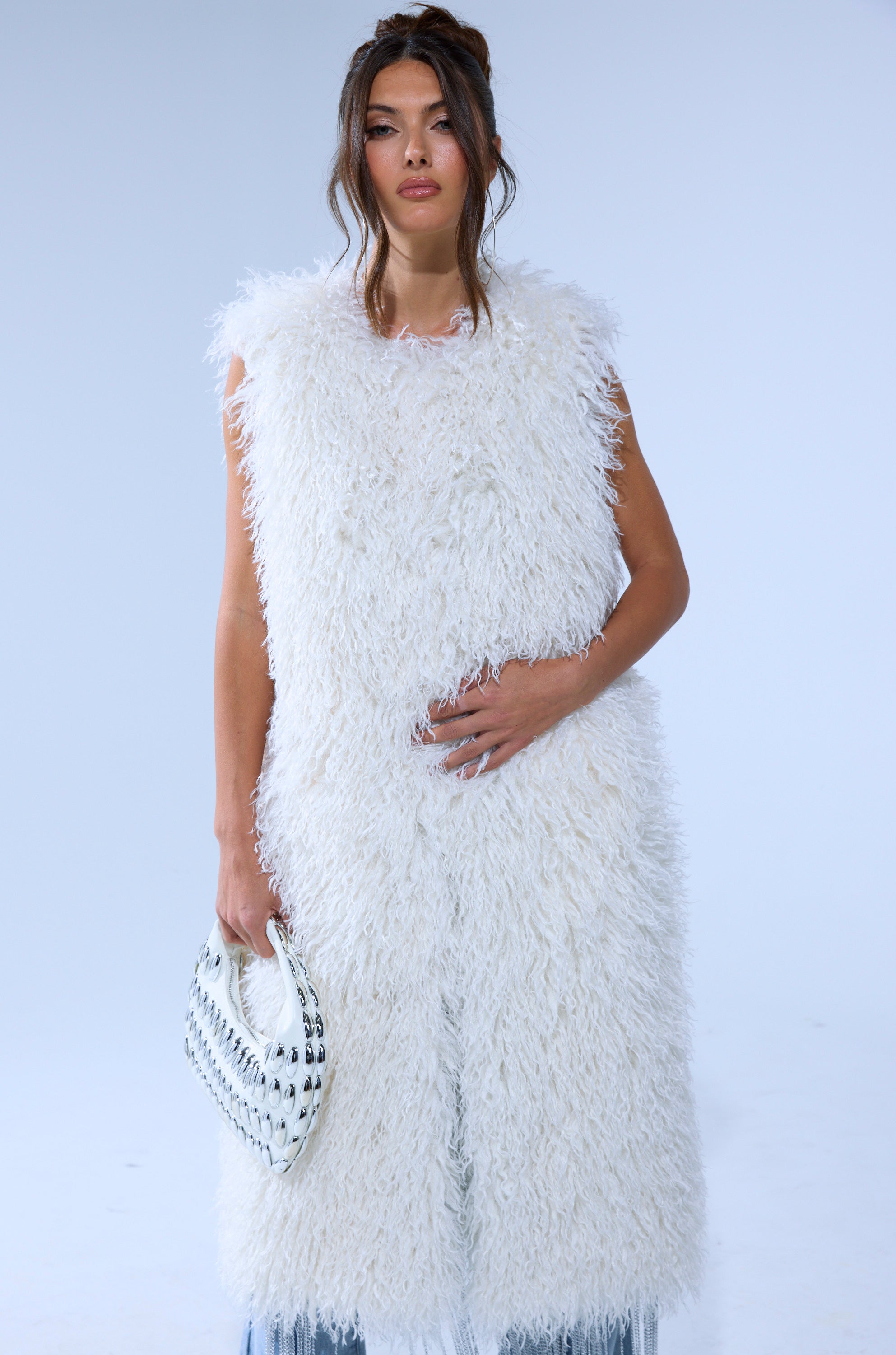 A woman in the ANOTHER LANE MOHAIR VEST holds a textured white clutch in her left hand, standing before a plain light background with loose wavy hair.