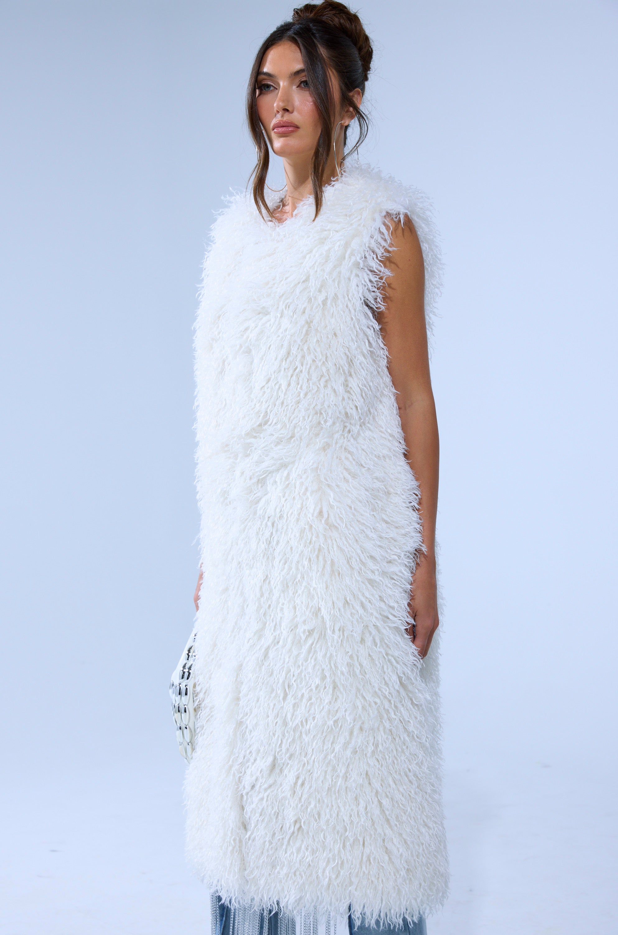 A woman with long brown hair in an updo wears the ANOTHER LANE MOHAIR VEST, a sleeveless, long white faux fur vest. She holds a small white bag and stands against a plain, light blue-gray background.