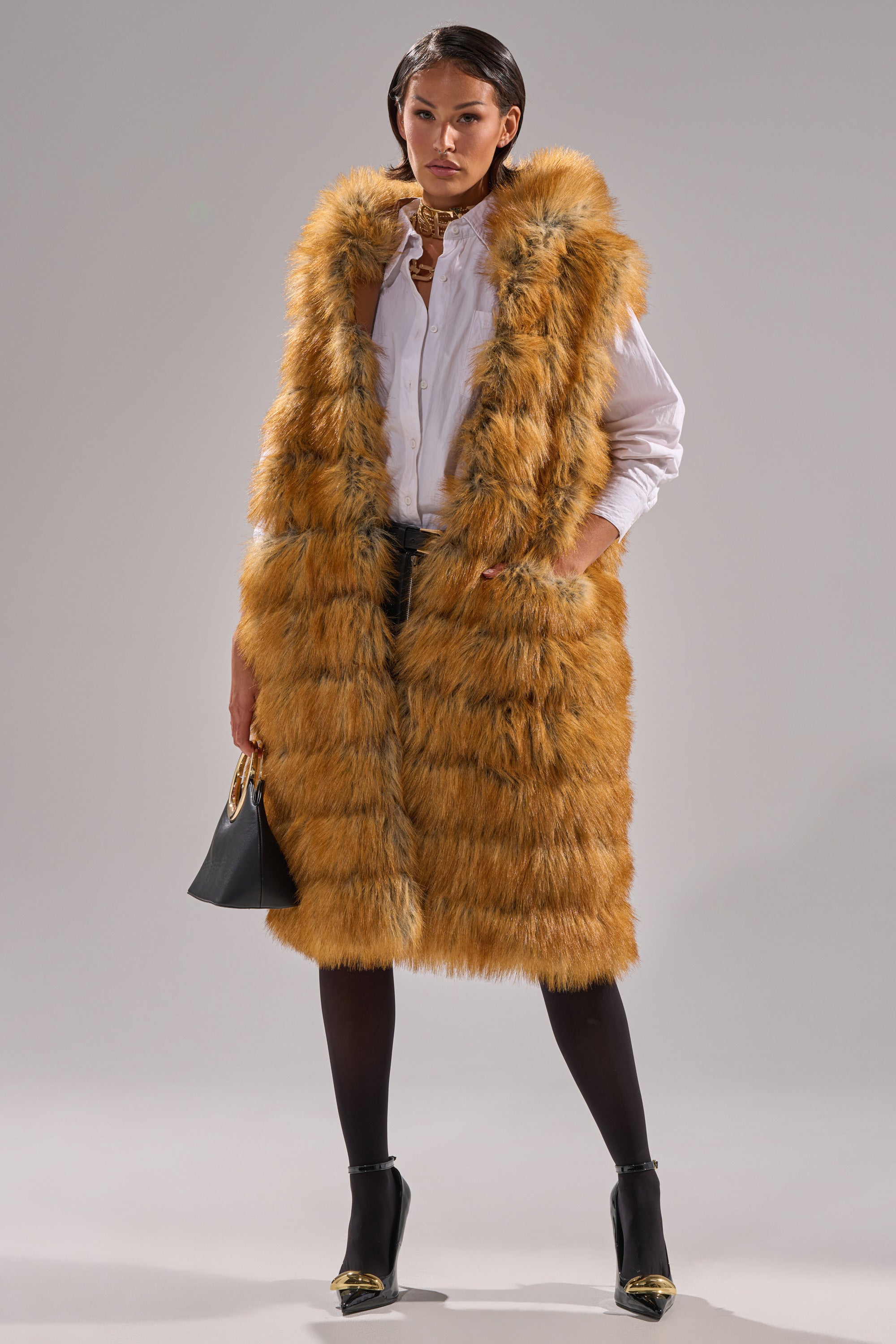 A woman stands confidently in the SLY HOODED FAUX FUR VEST, a longline sleeveless orange piece layered over a white shirt and dark skirt. She accessorizes with a black handbag, black tights, and heels against a plain light background.