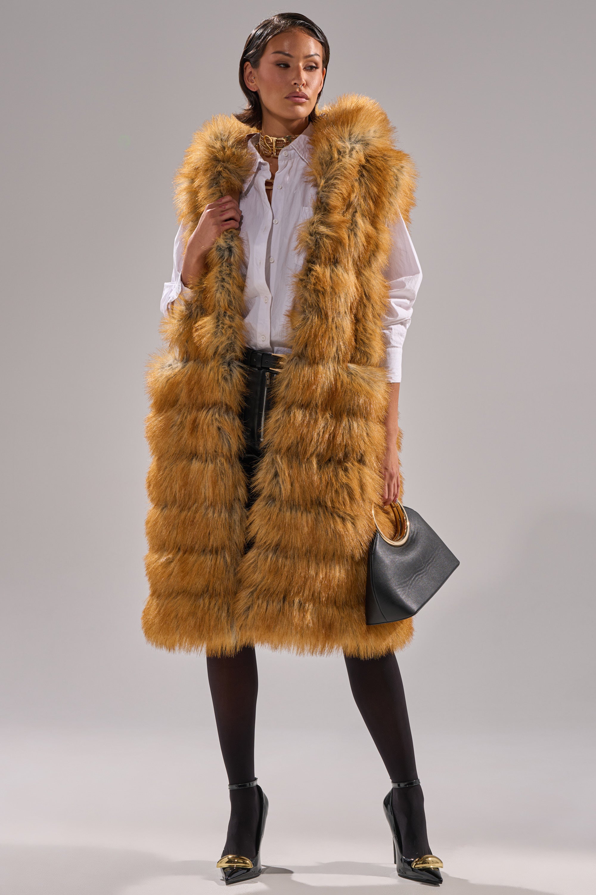 A woman stands confidently against a gray background wearing the SLY HOODED FAUX FUR VEST in orange-brown over a white shirt, black skirt, black tights, gold heels, and holding a black handbag.