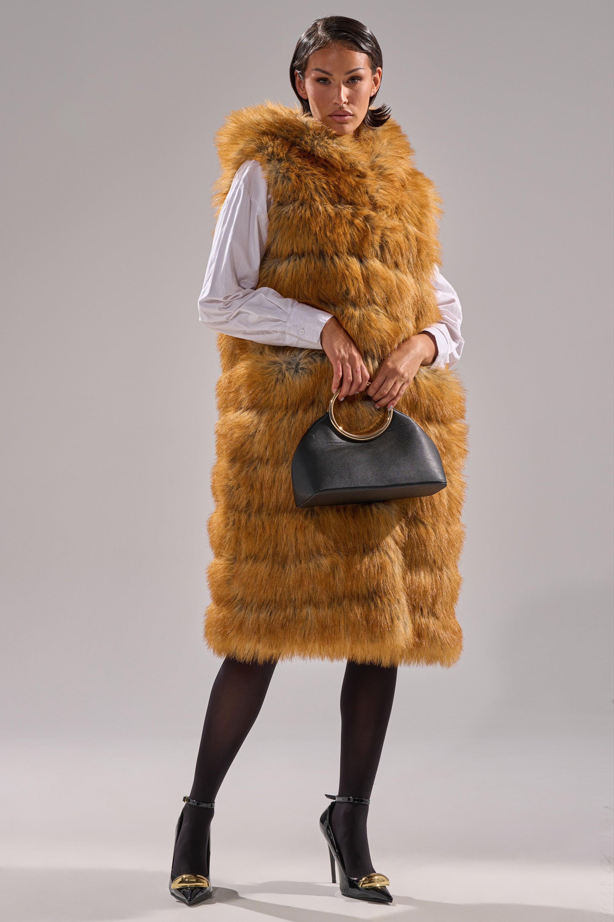 A woman stands against a plain background wearing the SLY HOODED FAUX FUR VEST, a long, fluffy, brown faux fur piece over a white shirt. She accessorizes with a black handbag with a gold handle and black heels featuring gold tips.