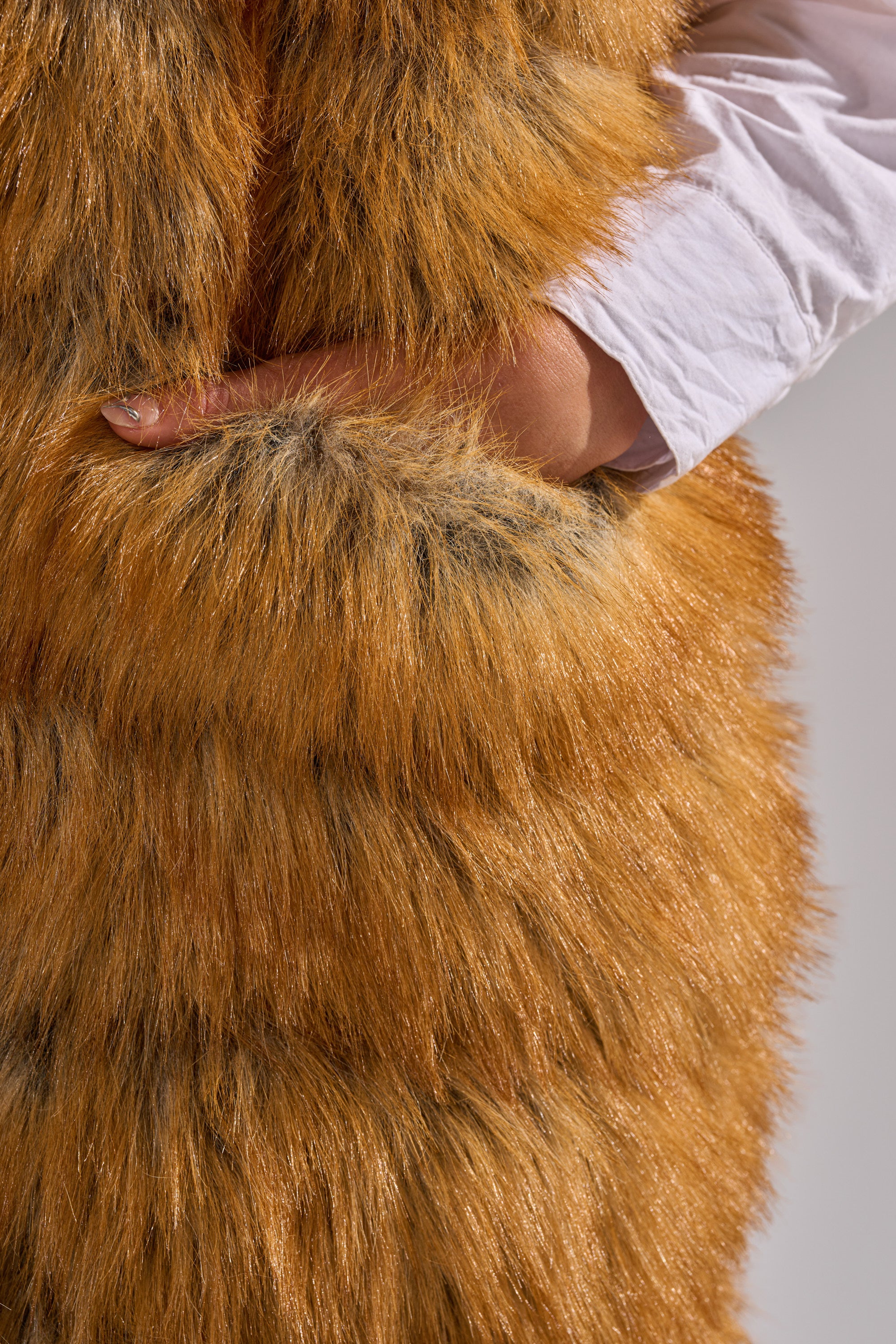 A person wearing a white long-sleeve shirt puts their hand in the pocket of the thick, brown SLY HOODED FAUX FUR VEST.