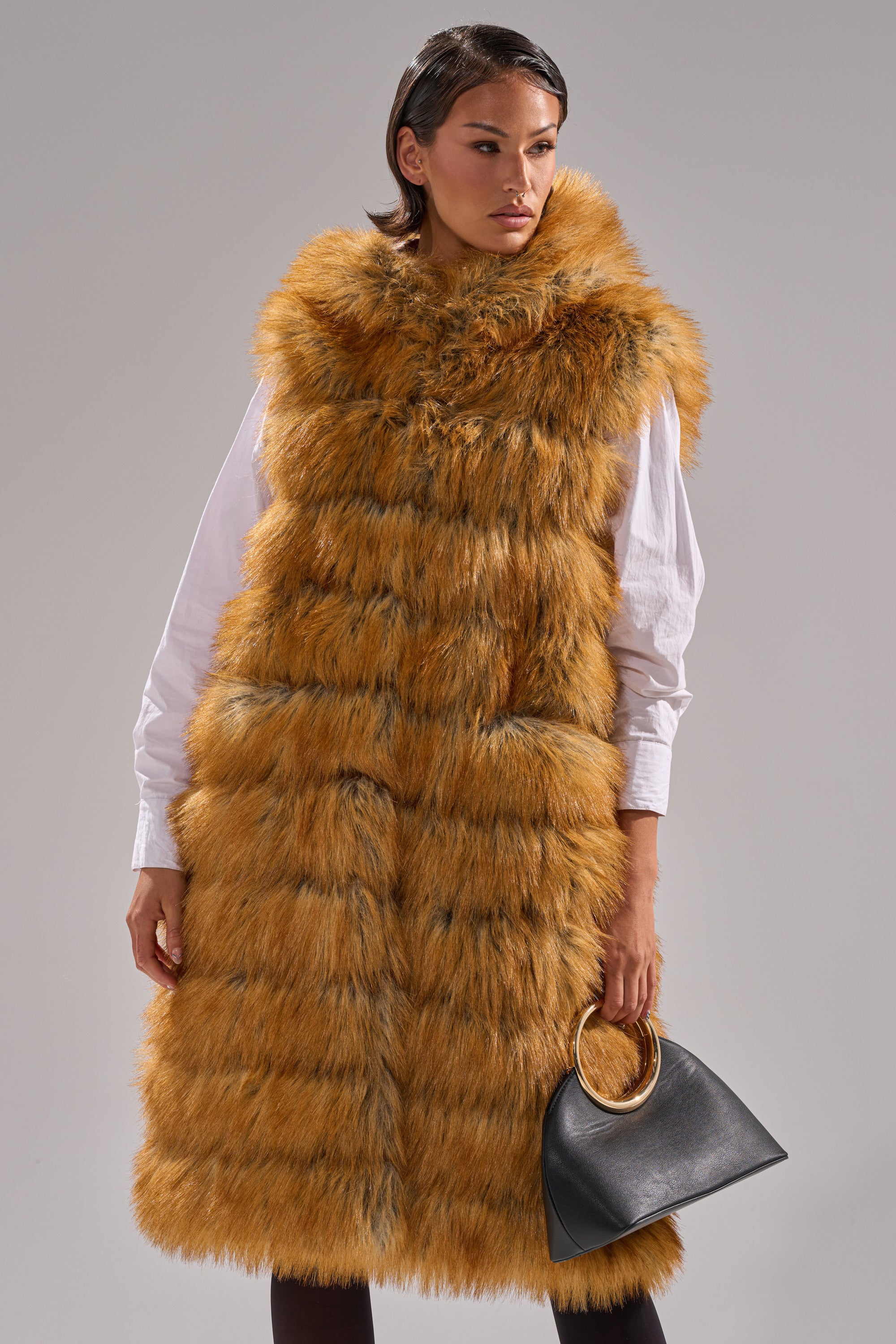 A woman in a SLY HOODED FAUX FUR VEST, longline and sleeveless in orange-brown, over a white shirt holds a black handbag with a gold handle while standing against a plain gray background.
