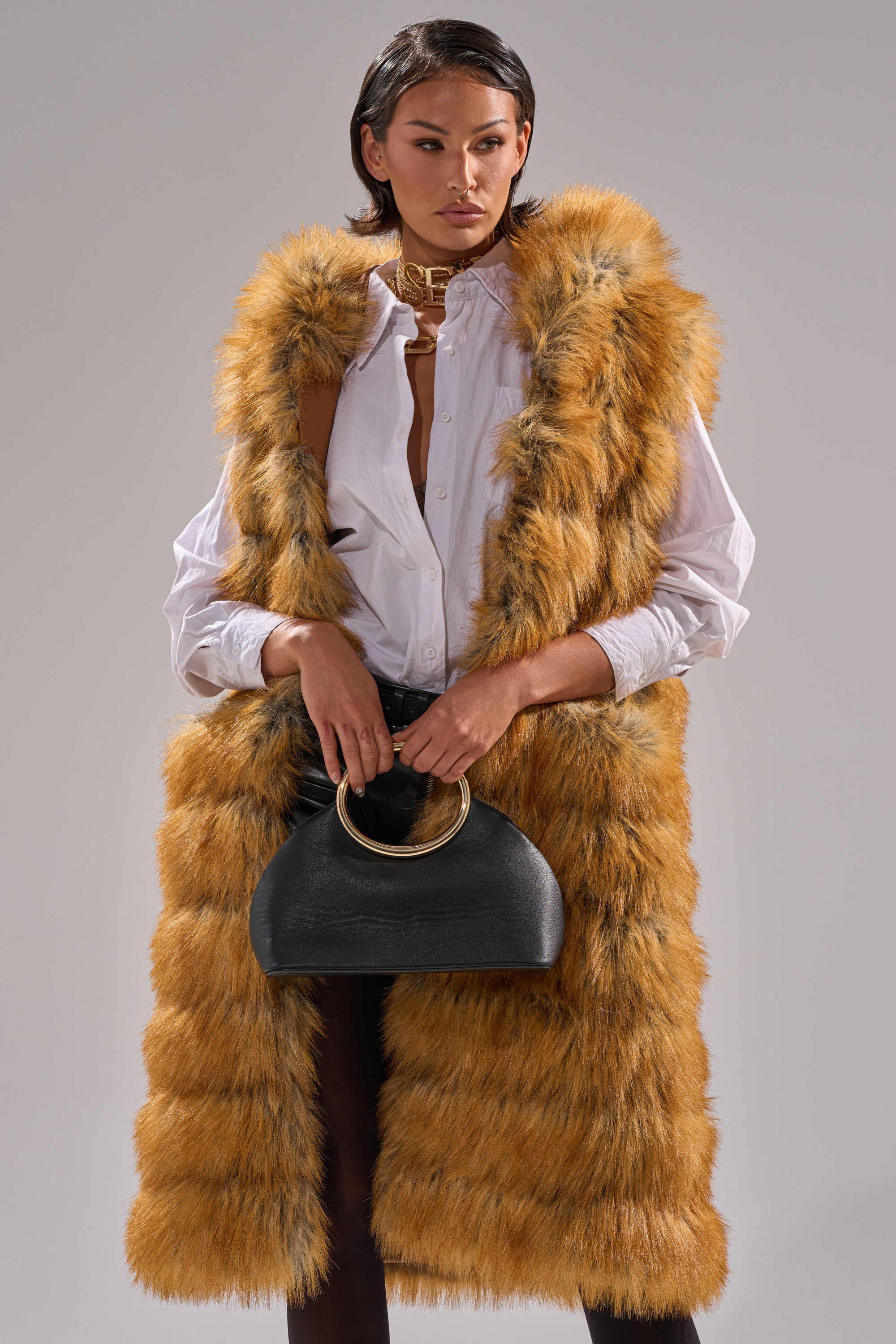 A woman with short dark hair and a confident expression wears the SLY HOODED FAUX FUR VEST over a white button-up shirt and black pants, holding a black handbag with a large gold ring handle.