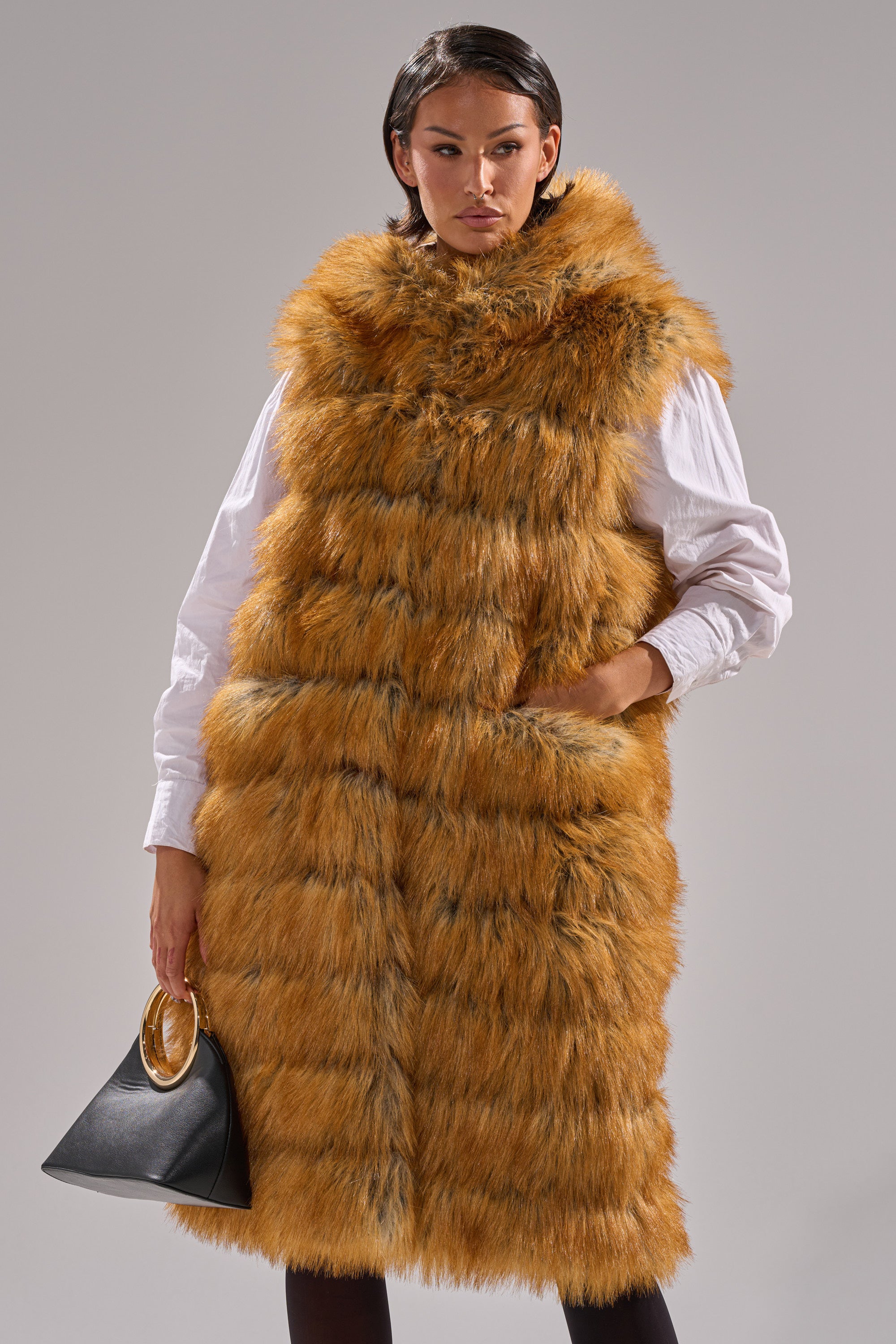 Wearing the SLY HOODED FAUX FUR VEST in orange-brown over a white shirt, a person holds a black handbag with a gold handle, standing against a plain gray background.