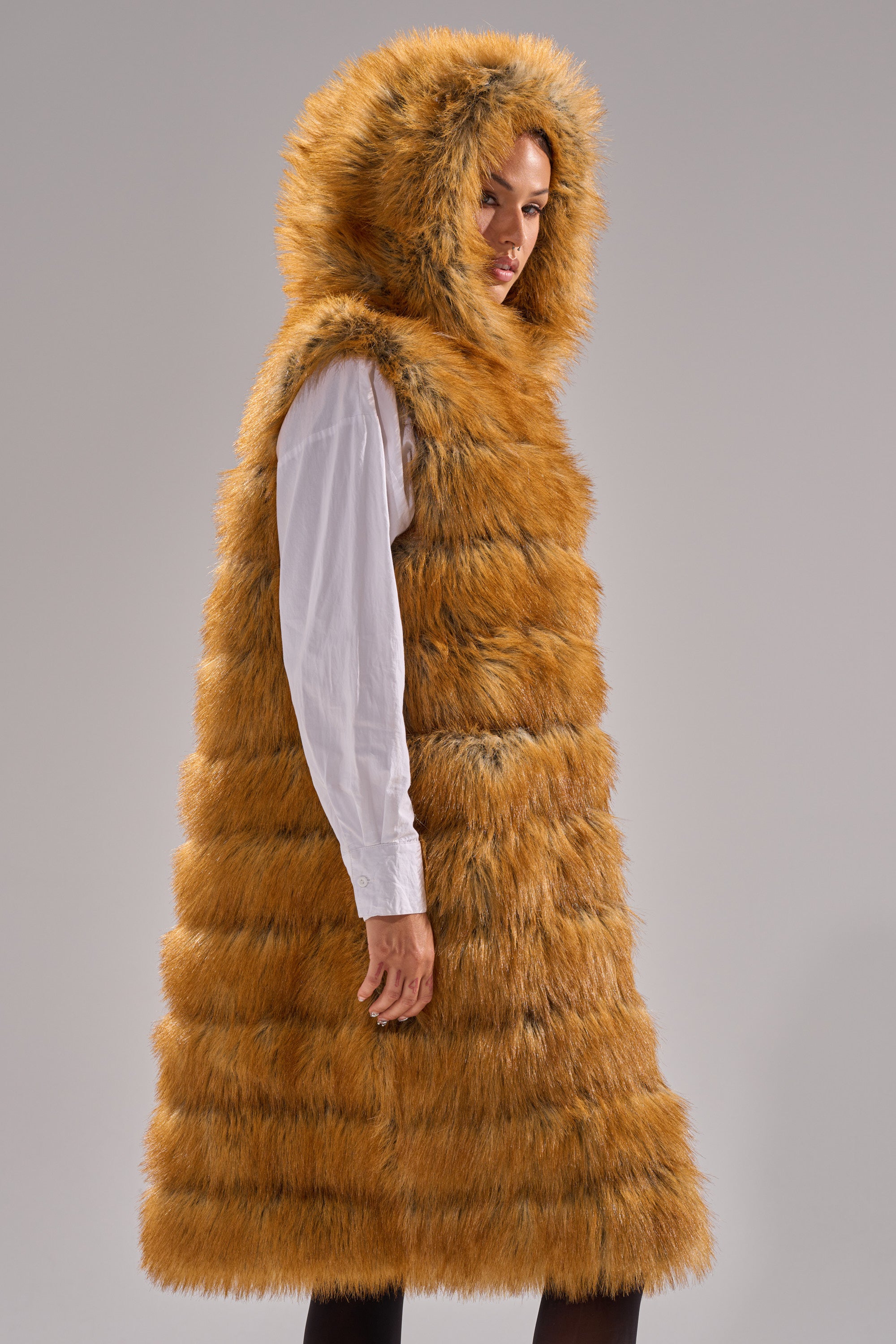 A person wears the SLY HOODED FAUX FUR VEST, a long reddish-brown hooded faux fur piece, over a white shirt while standing against a plain gray background.