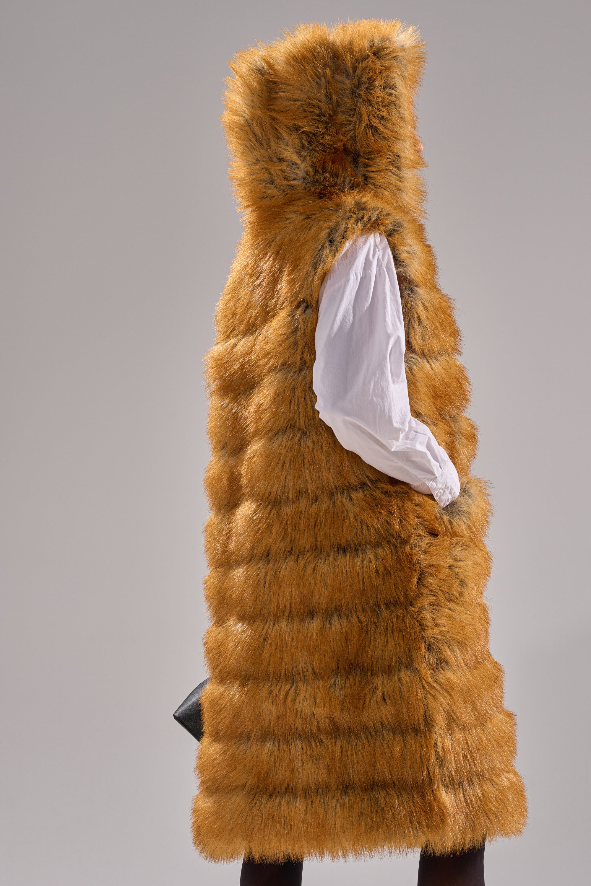 A person wears the SLY HOODED FAUX FUR VEST in orange-brown, styled with a white shirt and black gloves, standing against a plain gray background.