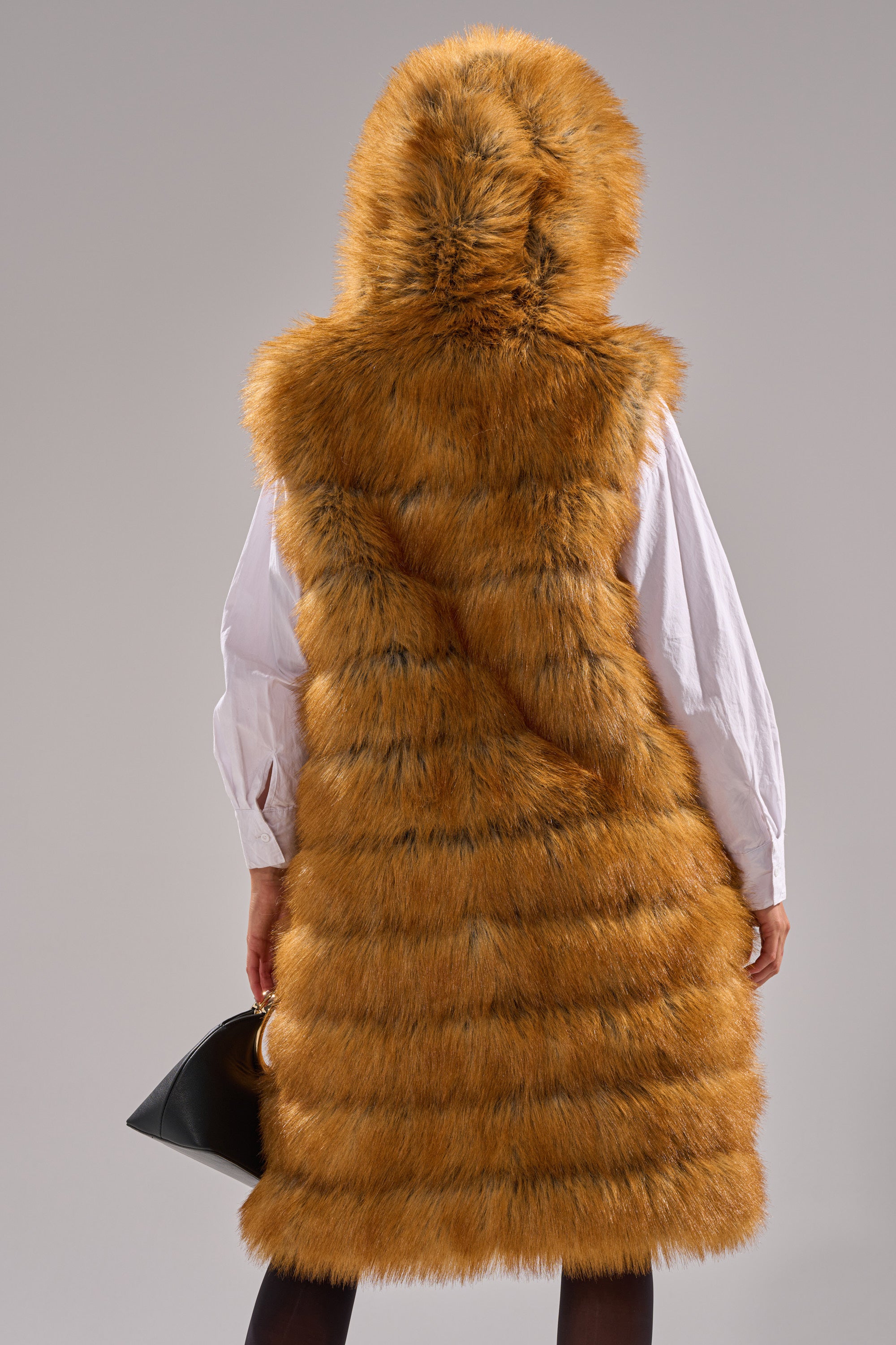 A person is seen from the back wearing the SLY HOODED FAUX FUR VEST, featuring orange and black stripes, over a white shirt and holding a black handbag against a plain gray background.