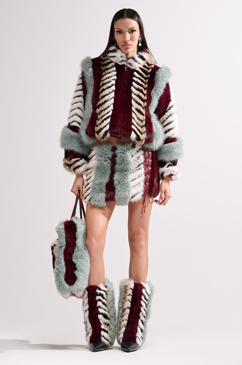 A woman showcases the HAVE IT ALL PANELED FUR SKIRT with a matching faux fur jacket, furry boots, and large furry bag in burgundy, white, black, tan, and pale blue, standing against a simple light background.
