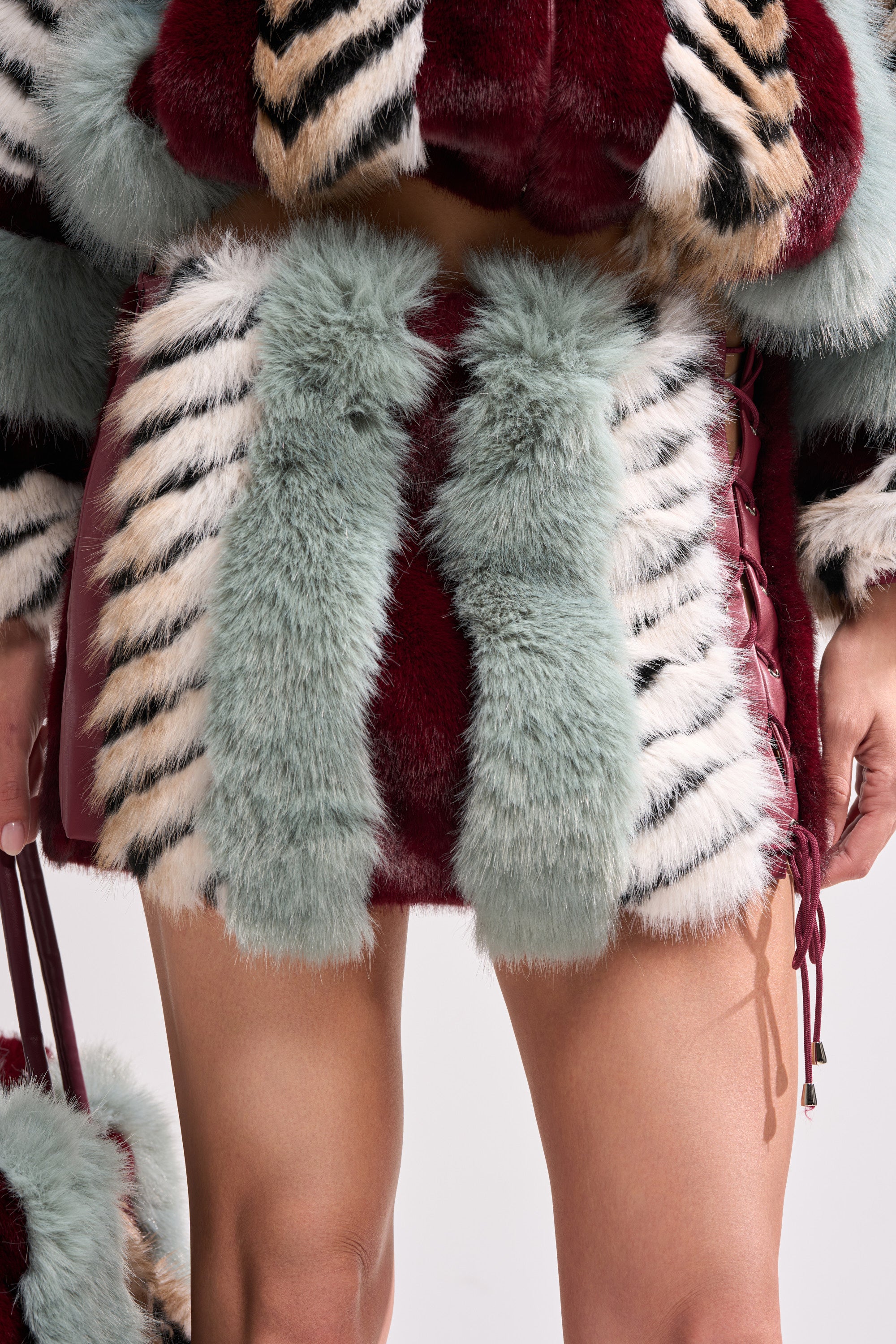 A person models the HAVE IT ALL PANELED FUR SKIRT, a short faux fur piece with vertical color block stripes in teal, burgundy, cream, and black. Their hands rest at their sides, showing bare legs beneath the skirt.