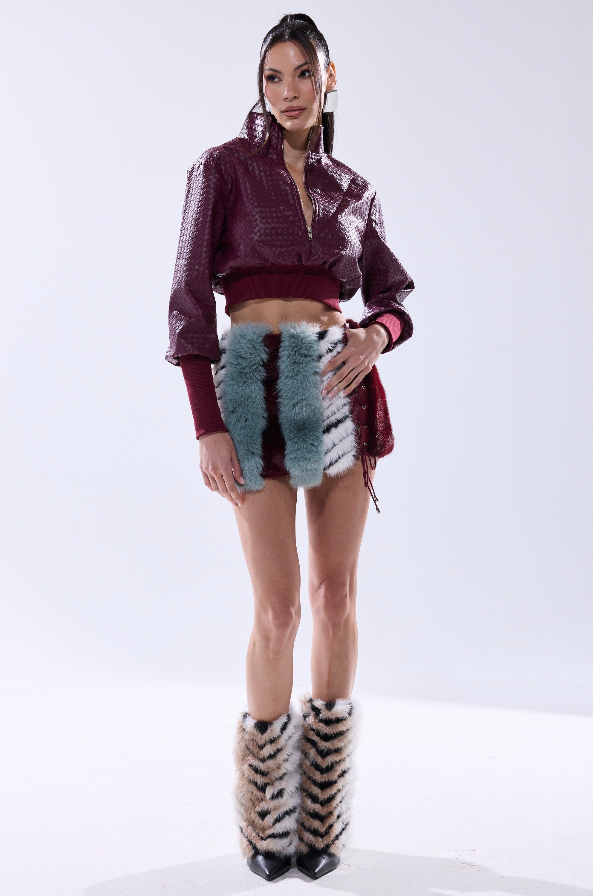 A woman stands against a plain background wearing the HAVE IT ALL PANELED FUR SKIRT, featuring fluffy vertical blue and white stripes, paired with a textured maroon jacket and matching zigzag-patterned furry boots.