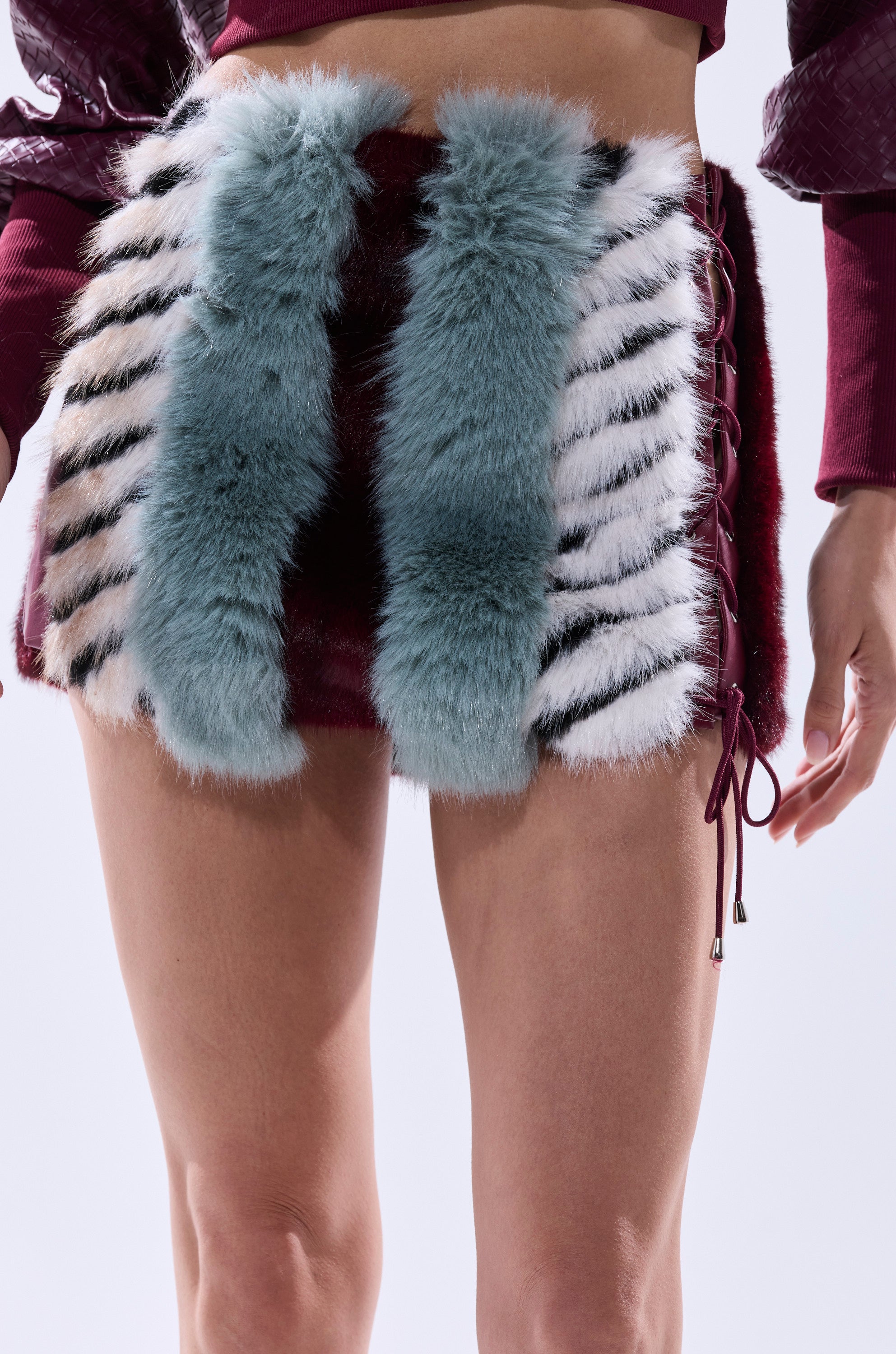 Someone is wearing the HAVE IT ALL PANELED FUR SKIRT, a short burgundy color block skirt with fluffy blue, white, and black faux fur stripes and side lace-up detail; their bare legs show.