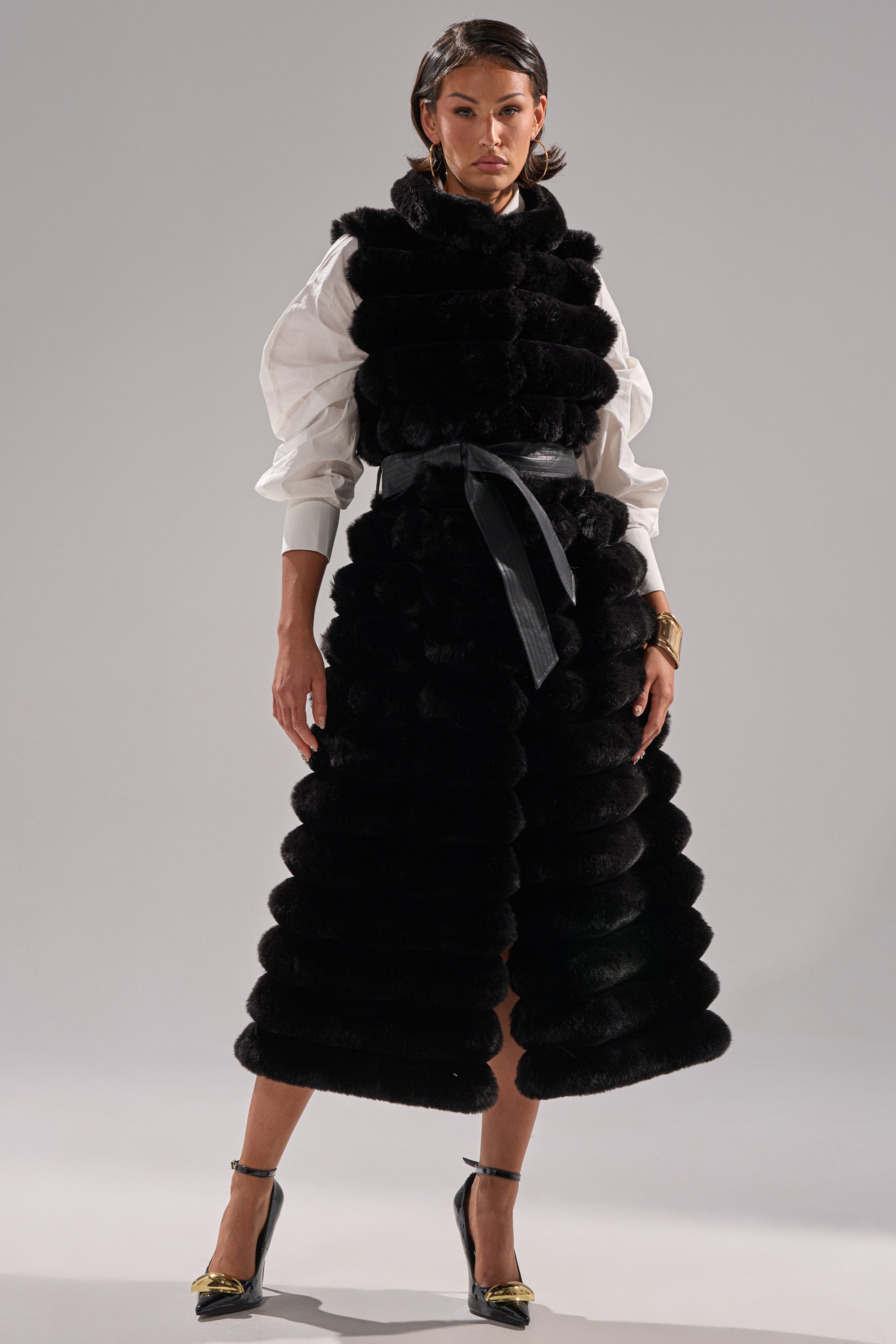 A woman stands confidently in the POPPY FAUX FUR VEST—a textured, sleeveless black faux fur vest over a white puff-sleeve blouse—belted at the waist, styled with black heels and gold anklets, against a neutral background.