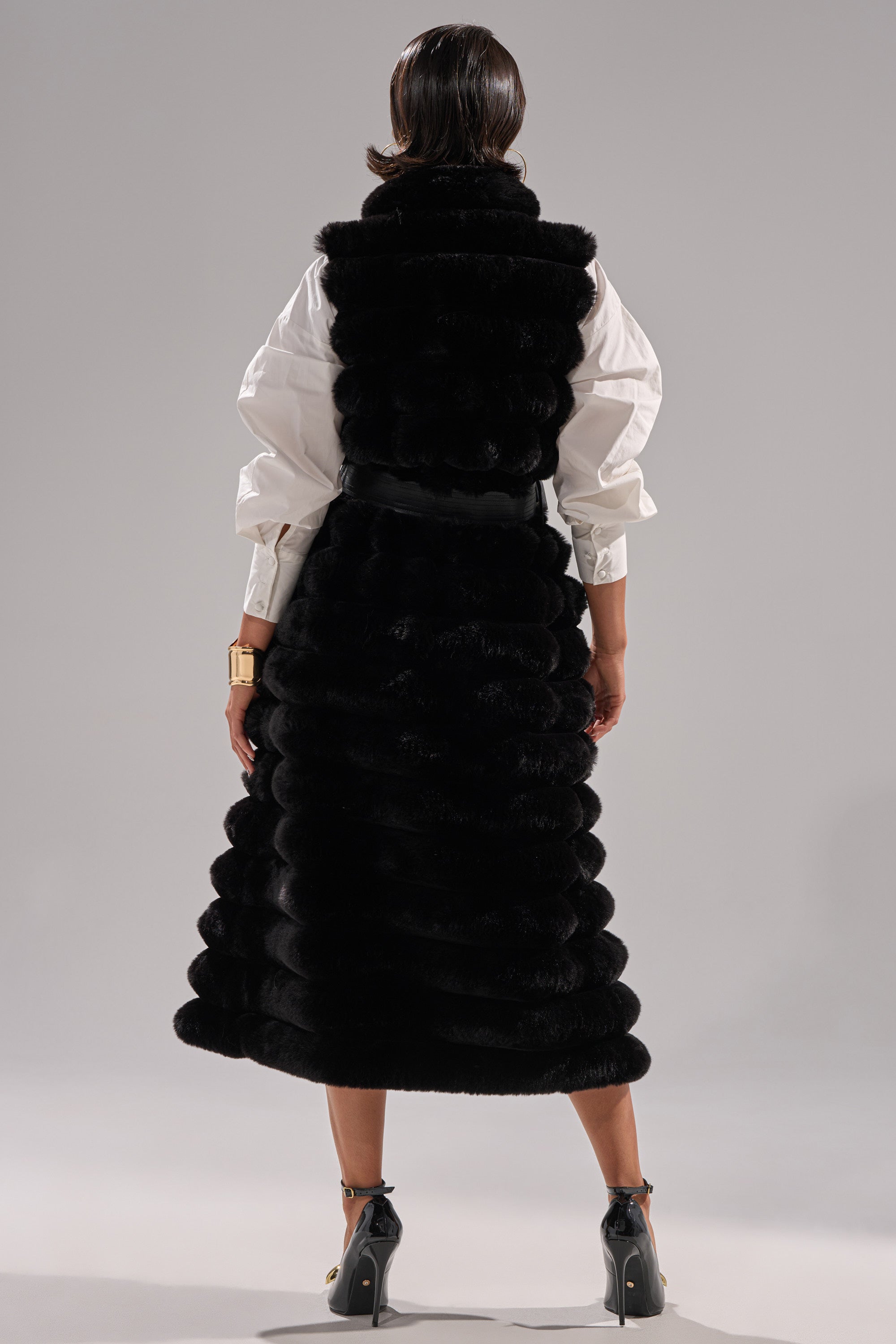 A person faces away from the camera in a POPPY FAUX FUR VEST, long and black with textured faux fur and a self-tie waist belt, layered over a white puff-sleeve shirt, black high heels, and a gold bracelet against a plain gray background.