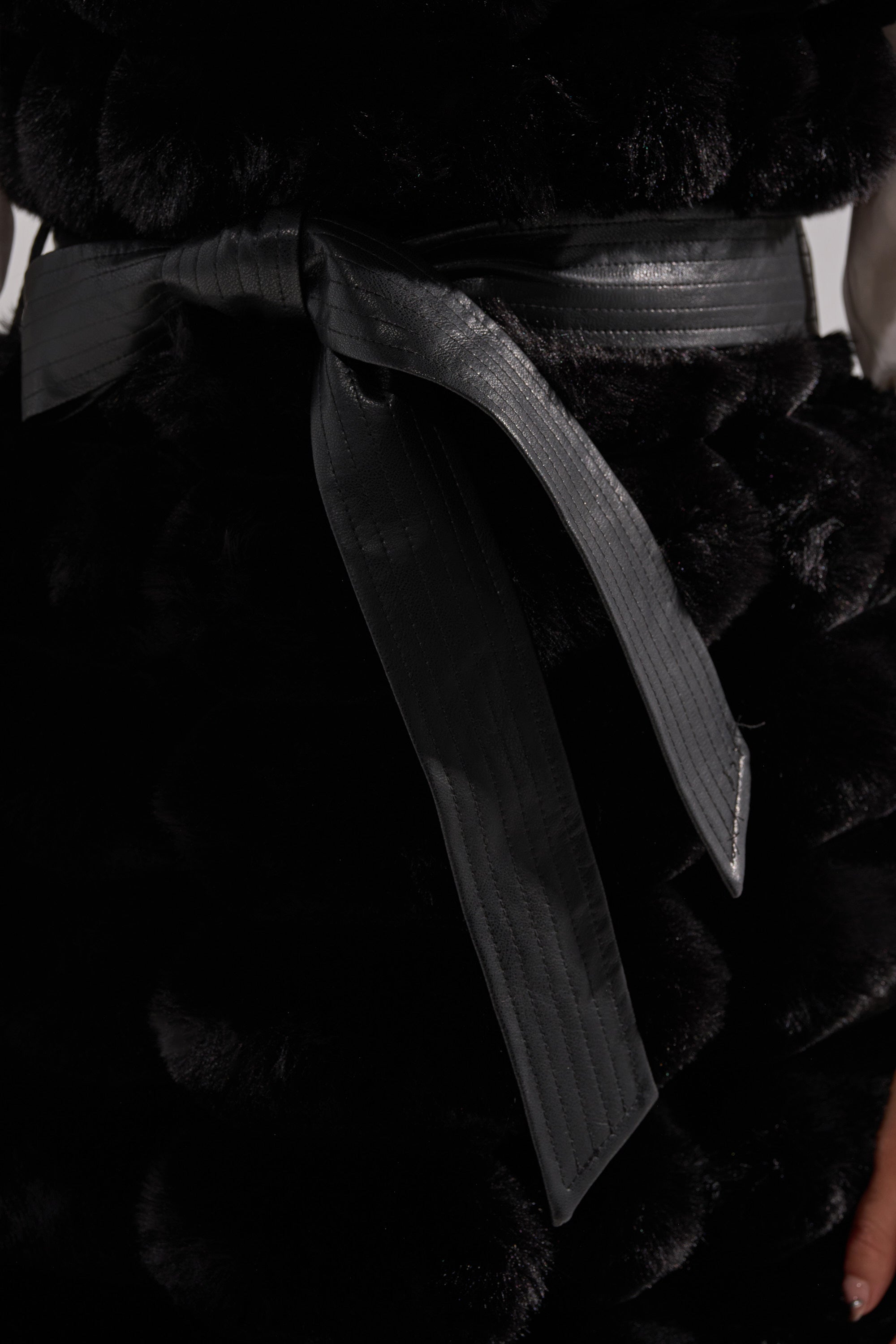 Close-up of the black POPPY FAUX FUR VEST, cinched at the waist with a self-tie belt and a hint of a white sleeve visible at the side.