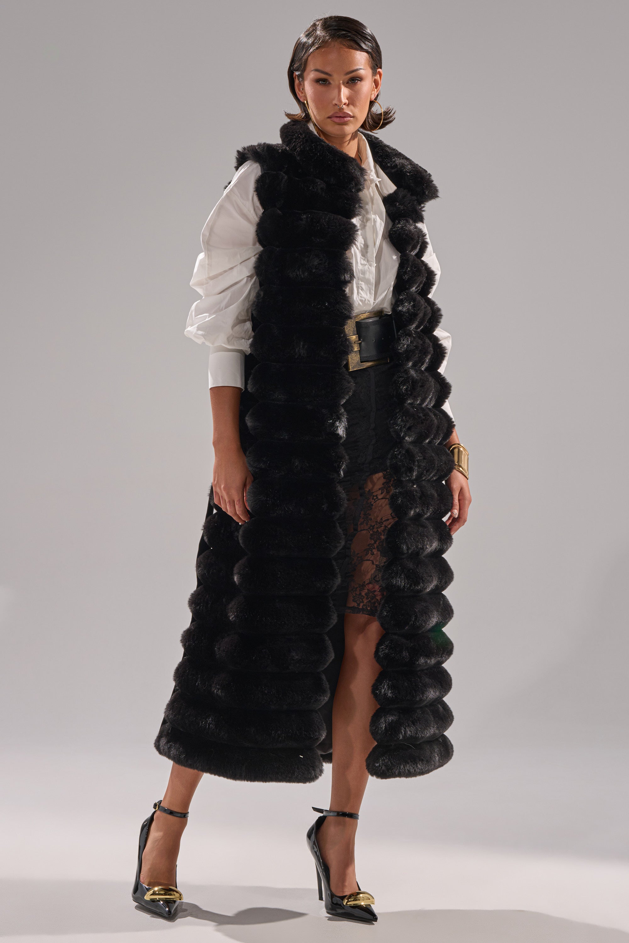 A woman with short dark hair and gold accessories stands confidently in the POPPY FAUX FUR VEST, featuring a longline cut and self-tie waist belt, layered over a white button-up shirt and black lace skirt with high heels.