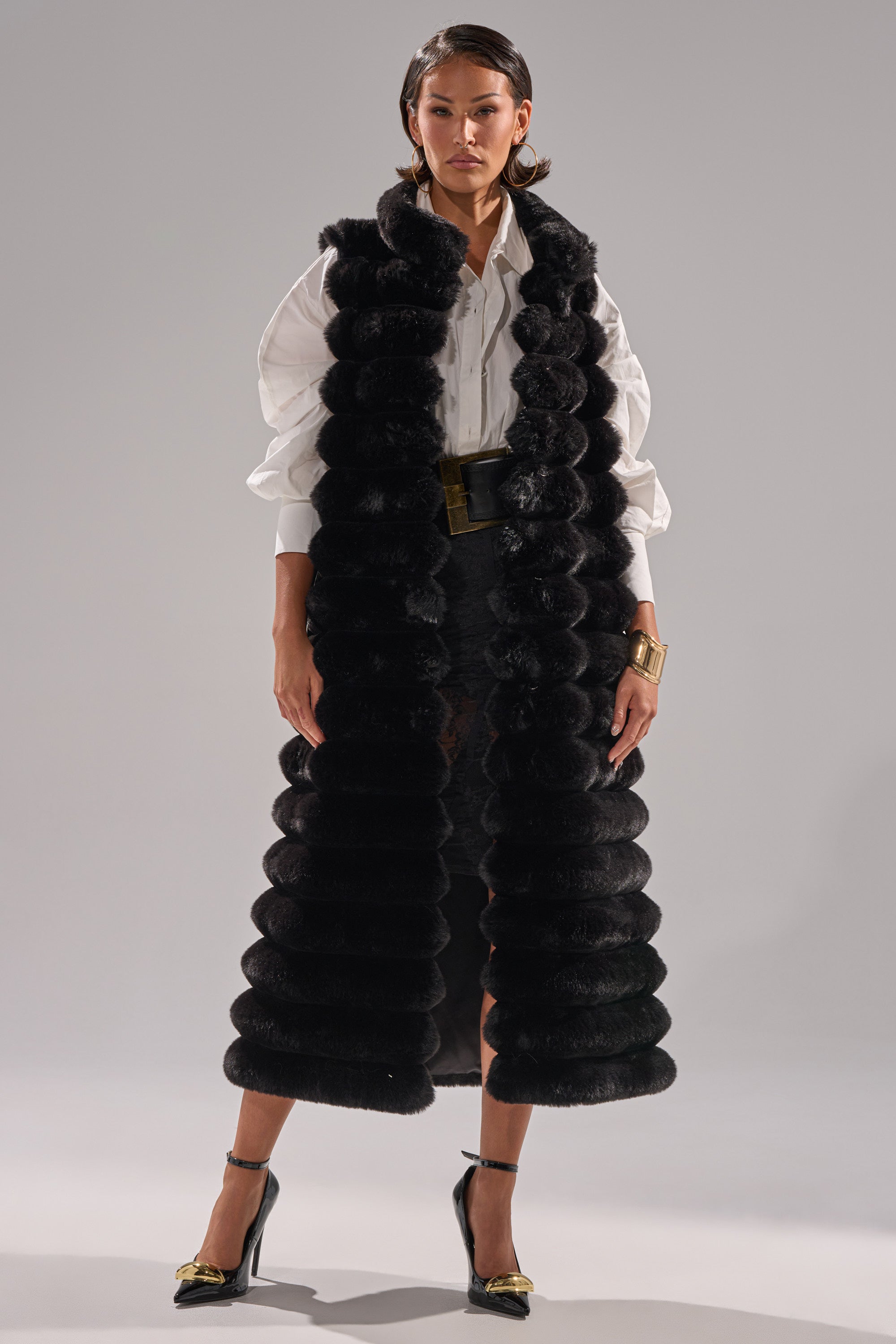 A woman stands facing forward wearing the POPPY FAUX FUR VEST— a longline black faux fur vest— layered over a white blouse with voluminous sleeves, a black skirt, self-tie waist belt, gold bracelet, and black high heels with gold accents.