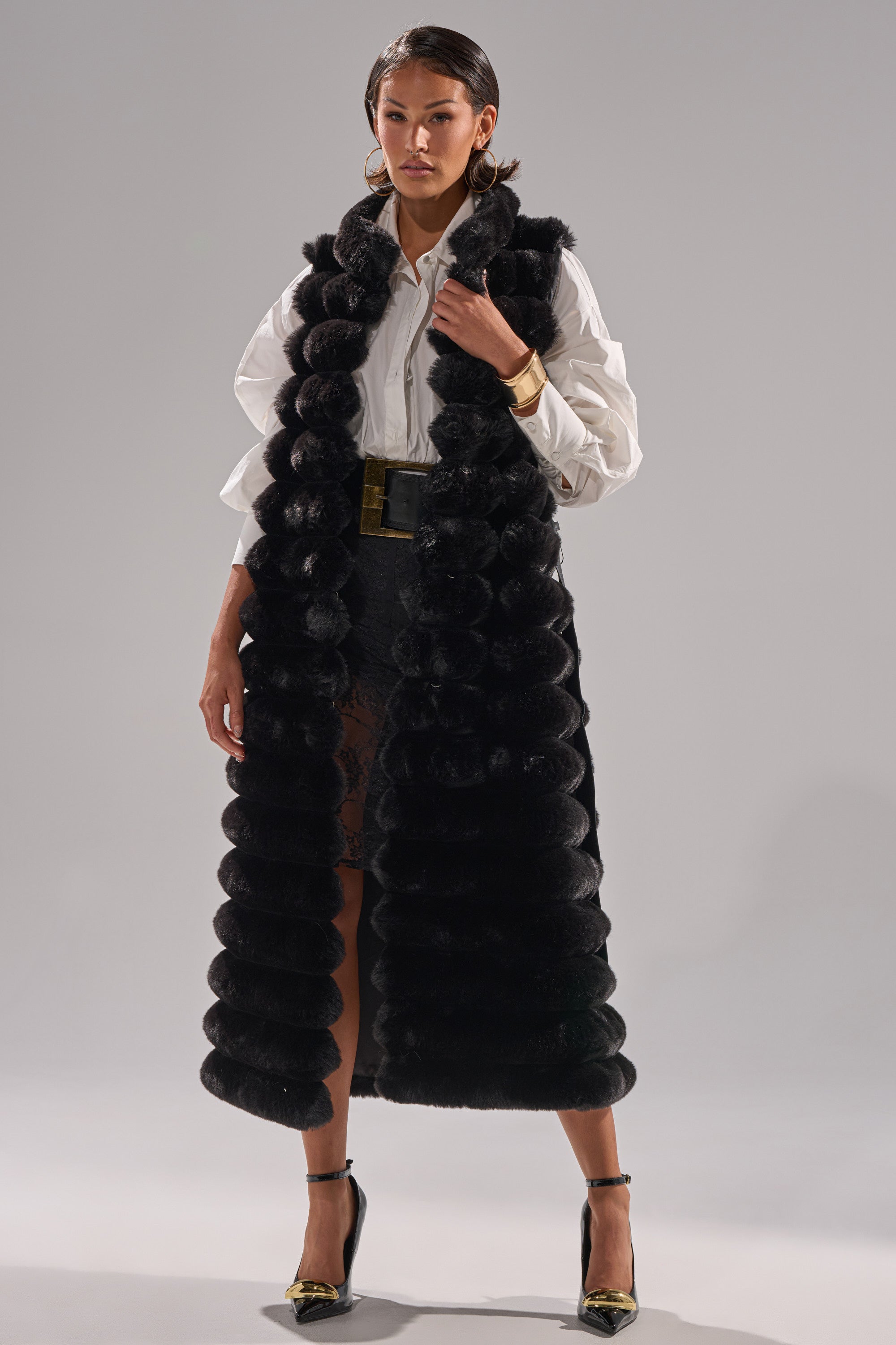 A woman poses confidently against a light background in the POPPY FAUX FUR VEST layered over a white blouse and black lace skirt, accessorized with gold hoop earrings, a wide belt, and black heels.