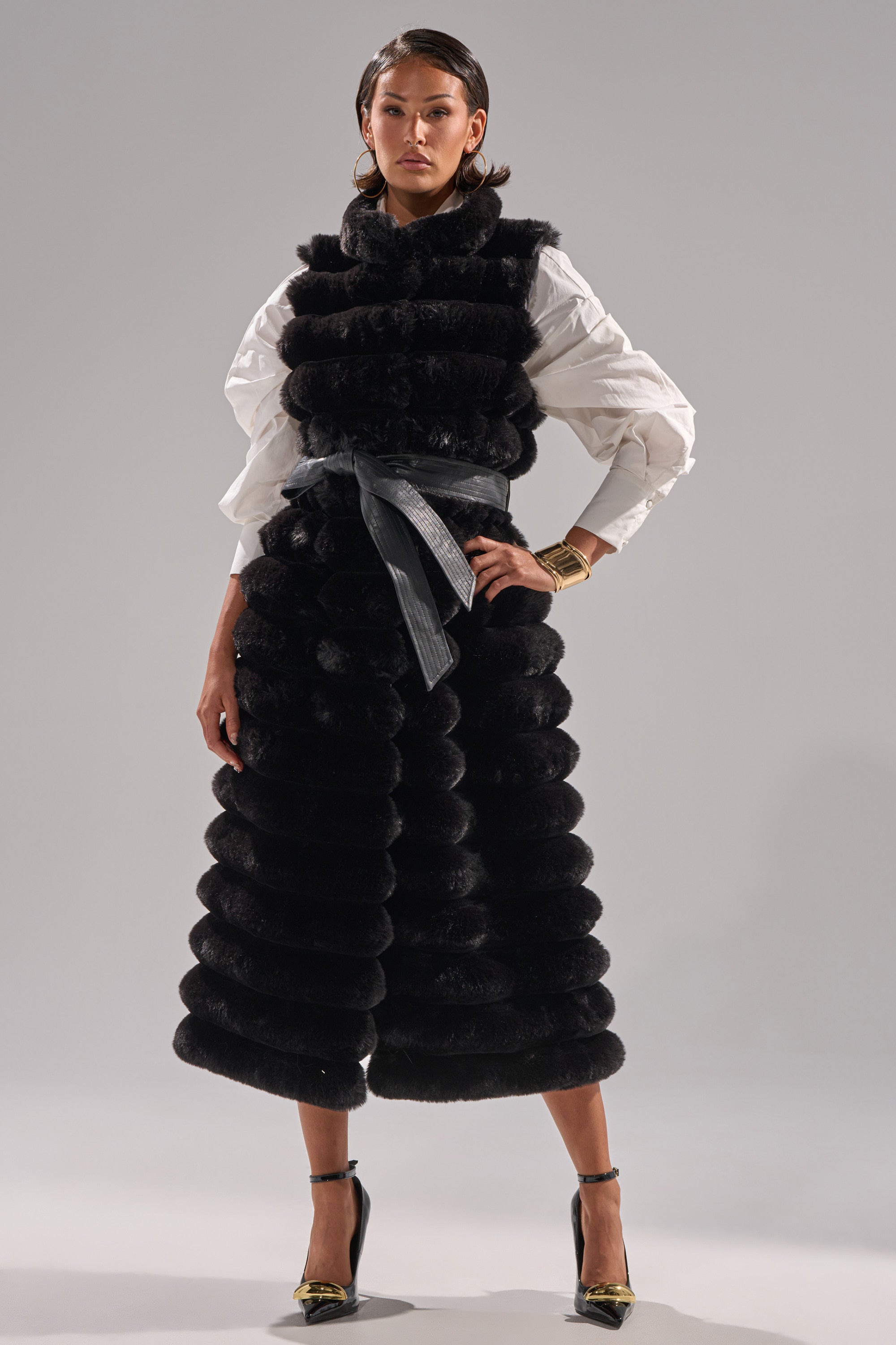 A woman confidently models the POPPY FAUX FUR VEST with matching wide-leg pants, a white blouse, black heels, a wide belt, and gold jewelry against a plain gray background.