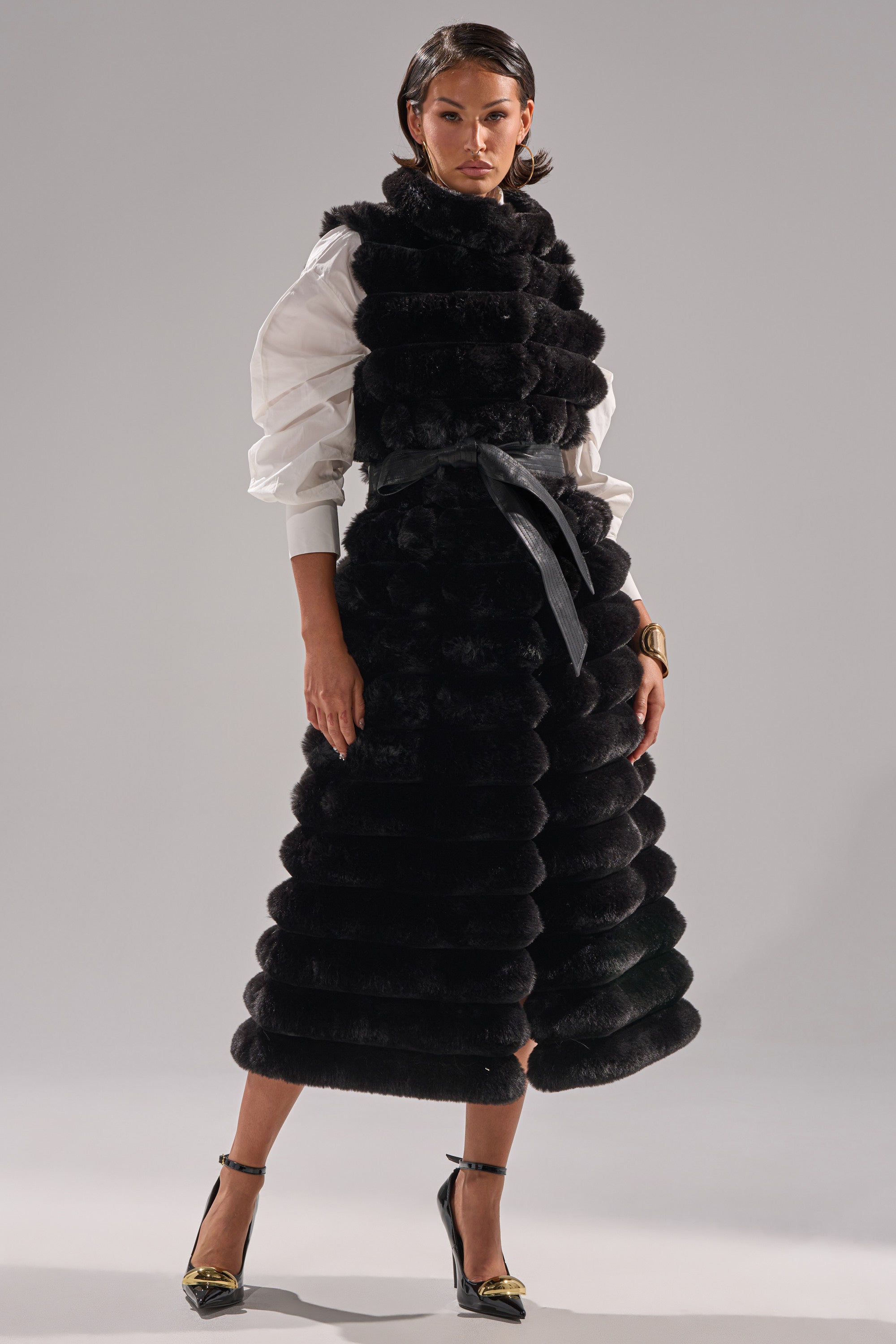 A woman stands confidently in the POPPY FAUX FUR VEST, cinched at the waist with a black belt, layered over a white blouse with puffed sleeves, and styled with black heels featuring gold accents and a gold bracelet.