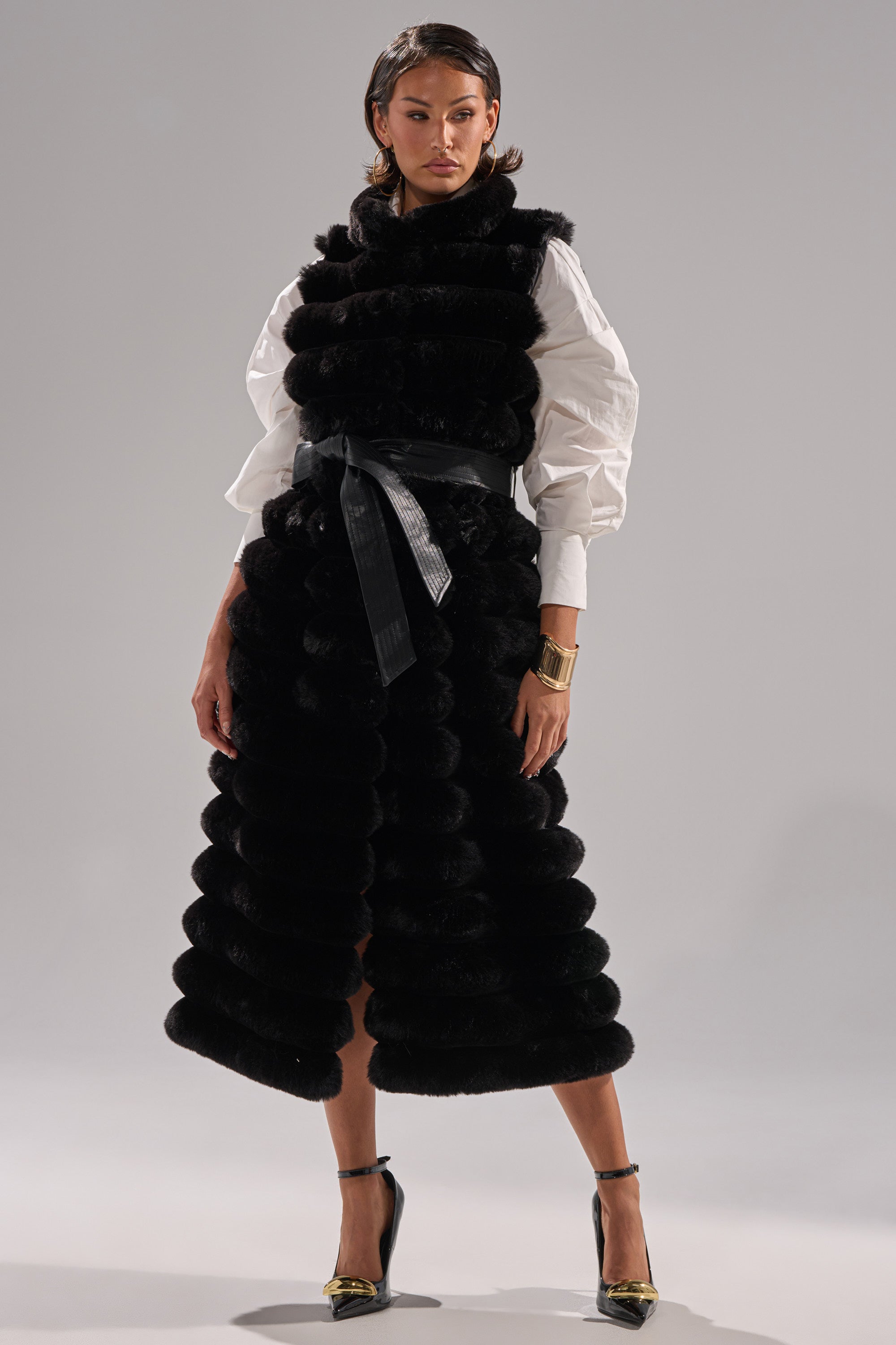 A woman poses in a white blouse with voluminous sleeves, the POPPY FAUX FUR VEST belted at the waist, black high heels with gold toes, and a gold cuff bracelet against a plain light gray background.