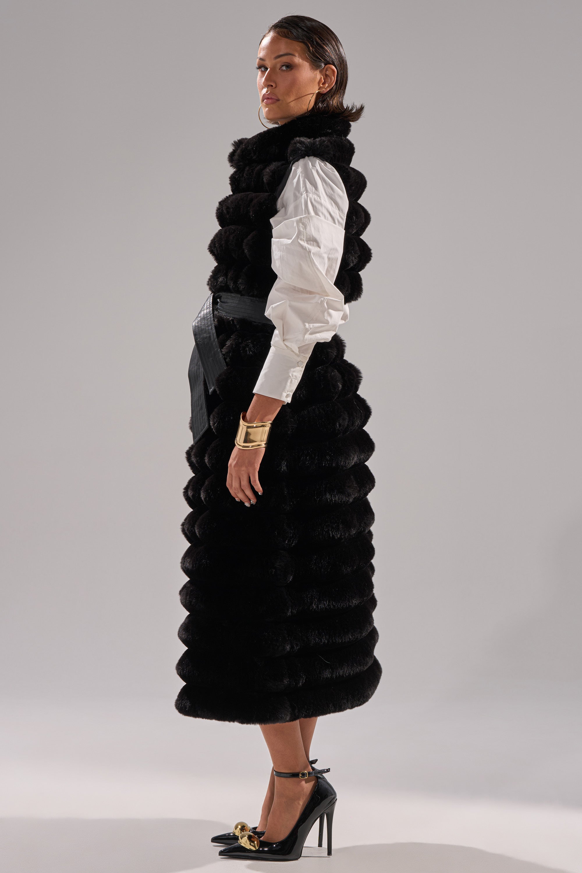 A woman stands sideways against a plain gray background, wearing the POPPY FAUX FUR VEST with a self-tie waist belt over a white shirt, black heels with gold accents, and gold bracelets.