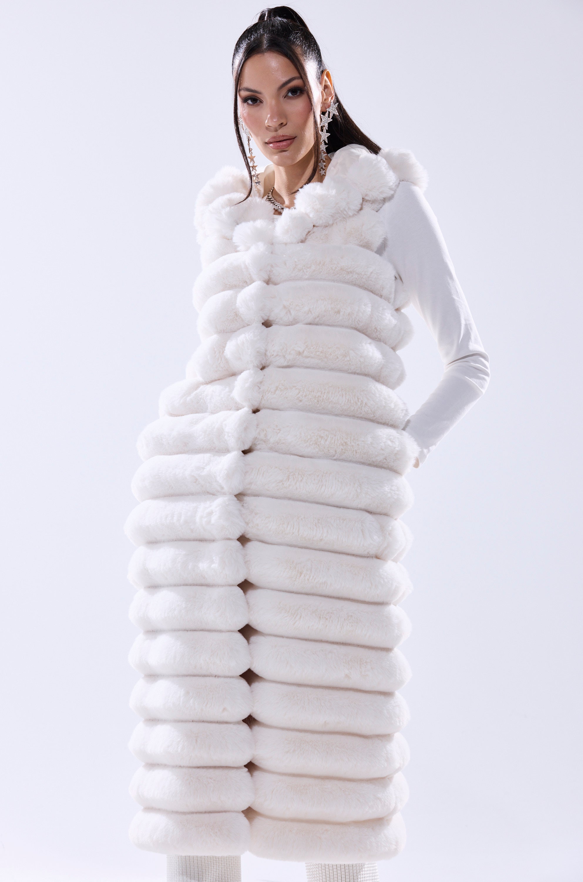 A woman models the POPPY FAUX FUR HOODED VEST—a long, plush white vest with horizontal ridges—over a white top and statement earrings, making this chic winter layer pop against a plain, light-colored background.