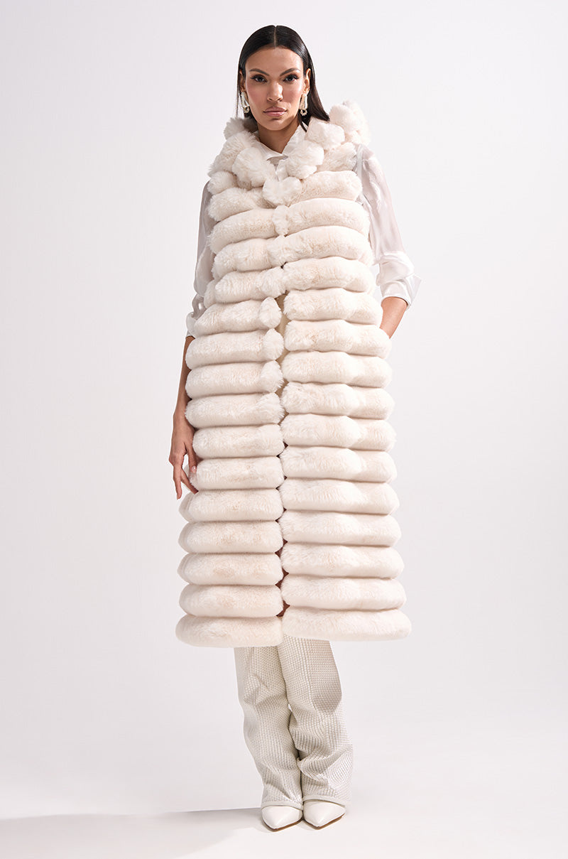 A woman models the POPPY FAUX FUR HOODED VEST—long, sleeveless, and white with horizontal padded sections—layered over a white blouse and pants, posed against a plain white background.