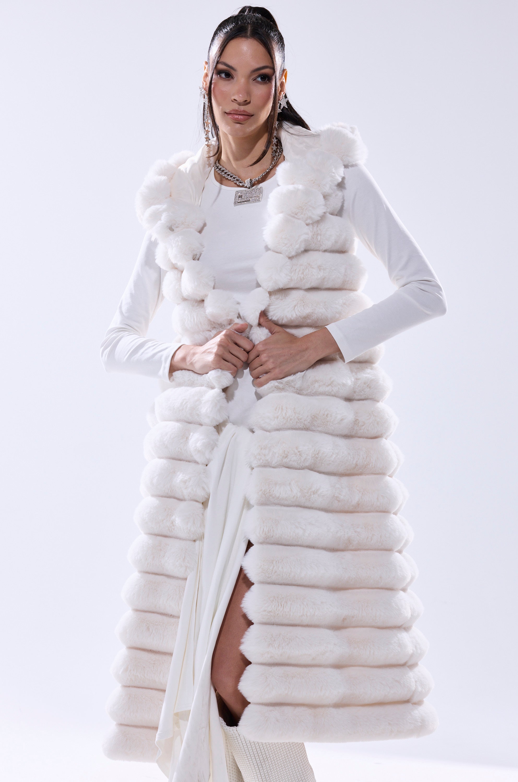 A woman models the POPPY FAUX FUR HOODED VEST, a longline sleeveless white piece with a hood, styled over a white dress. With straight dark hair and jewelry, she exudes confidence, making this vest an elegant winter layering choice.