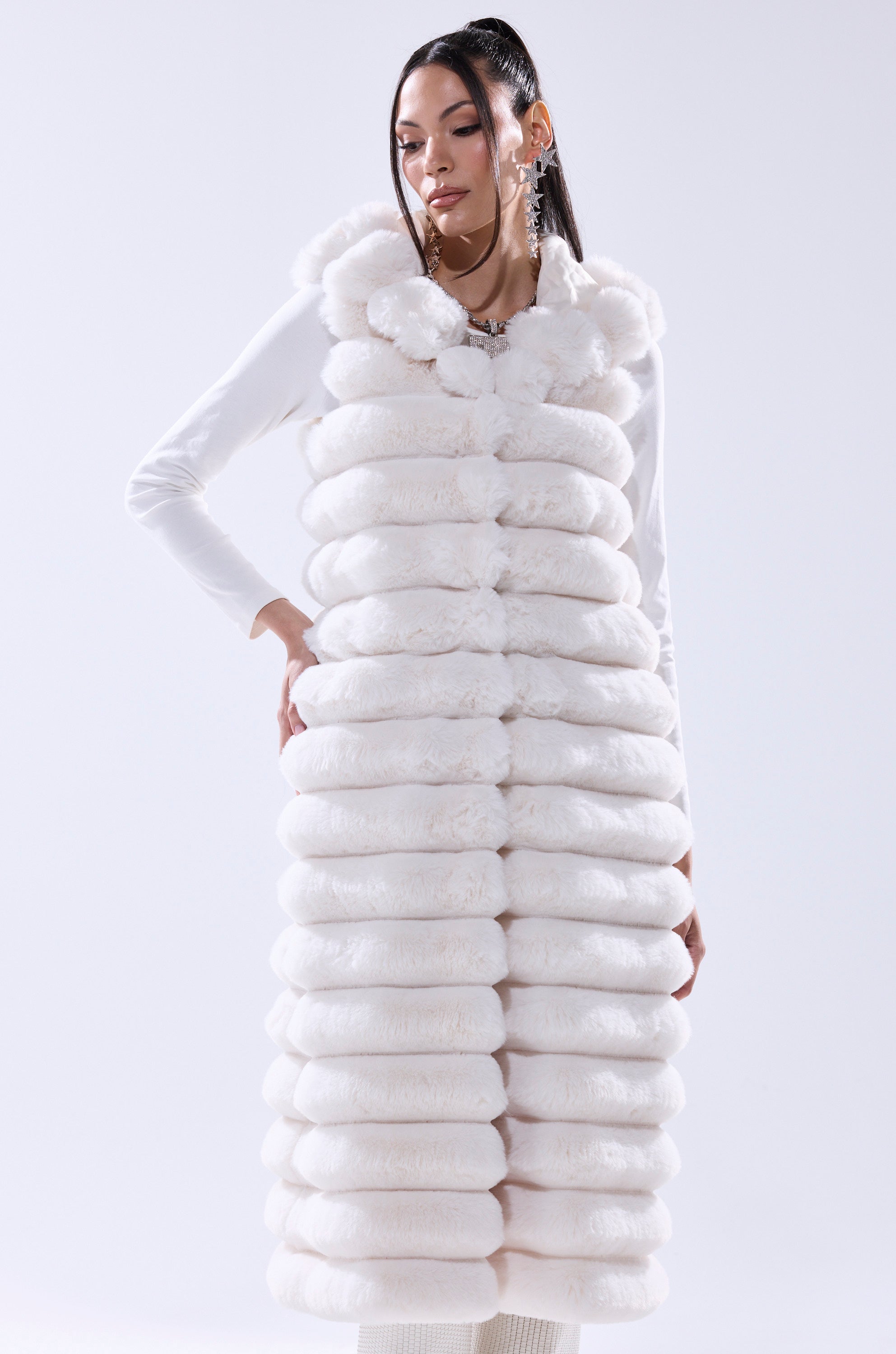A woman models the POPPY FAUX FUR HOODED VEST—a sleeveless, longline white vest with a large collar and hood—layered over a white long-sleeve top. A chic winter essential combining warmth and style against a light backdrop.
