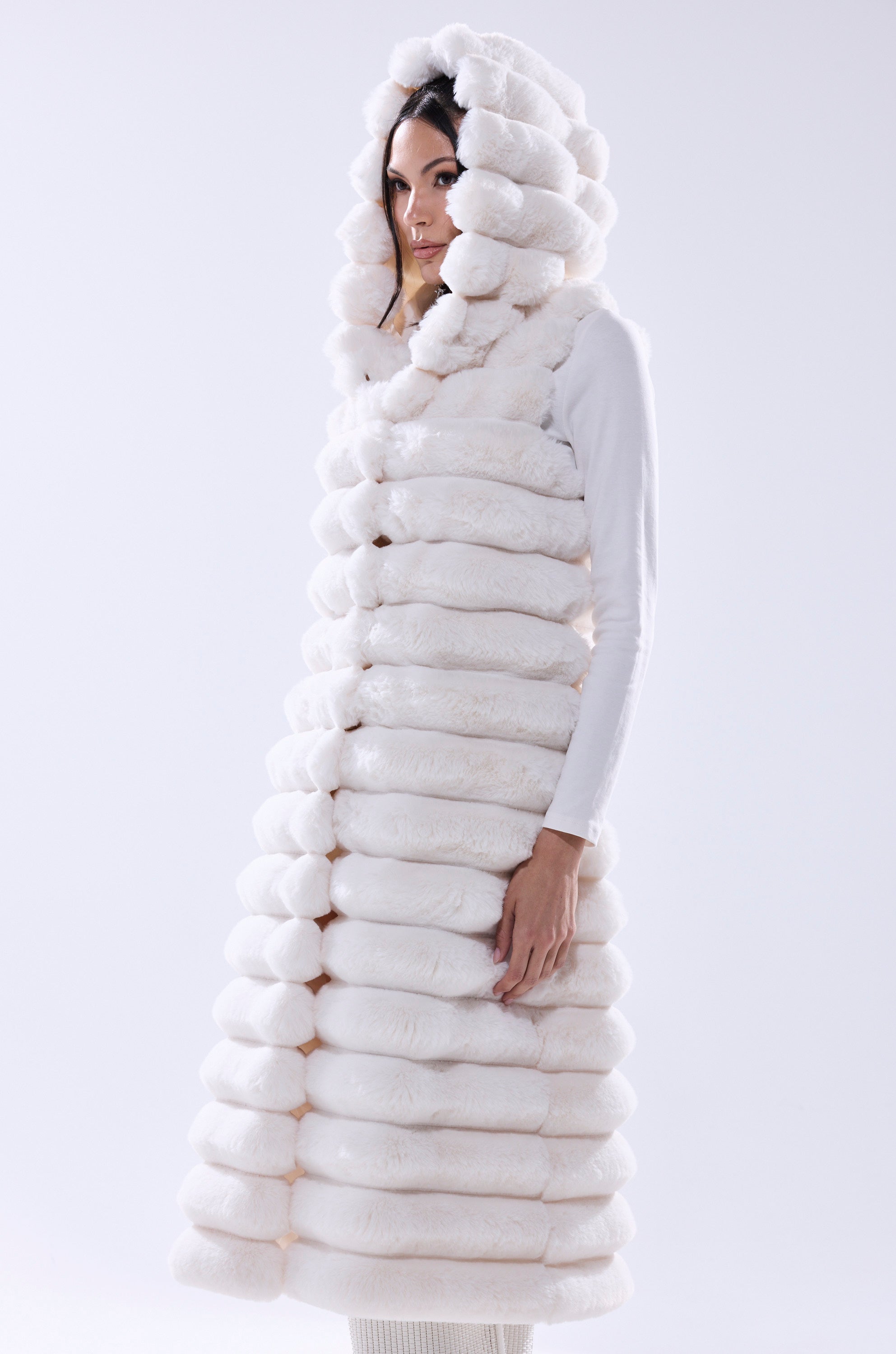 A woman models the POPPY FAUX FUR HOODED VEST, a chic, white, hooded longline layer with horizontal puffy sections—perfect for adding stylish warmth against a minimalist light background.