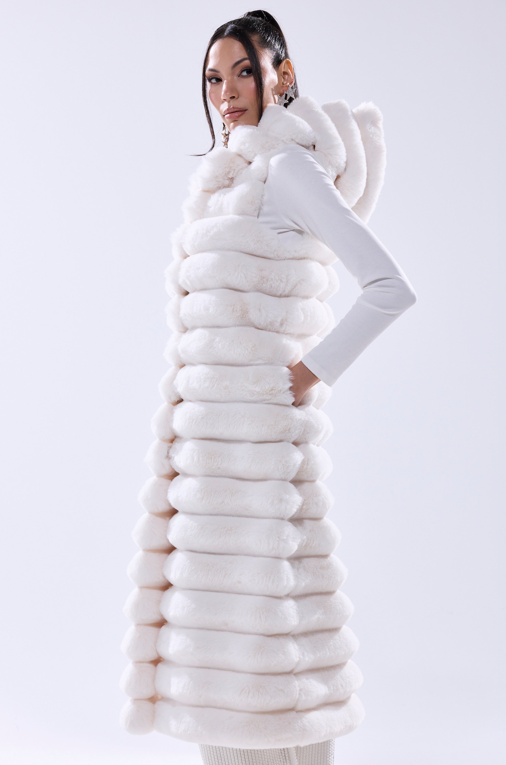 A woman confidently poses in the POPPY FAUX FUR HOODED VEST—a long, sleeveless white vest with a hood, high collar, and thick horizontal sections—against a plain light background, showcasing this chic winter layer.