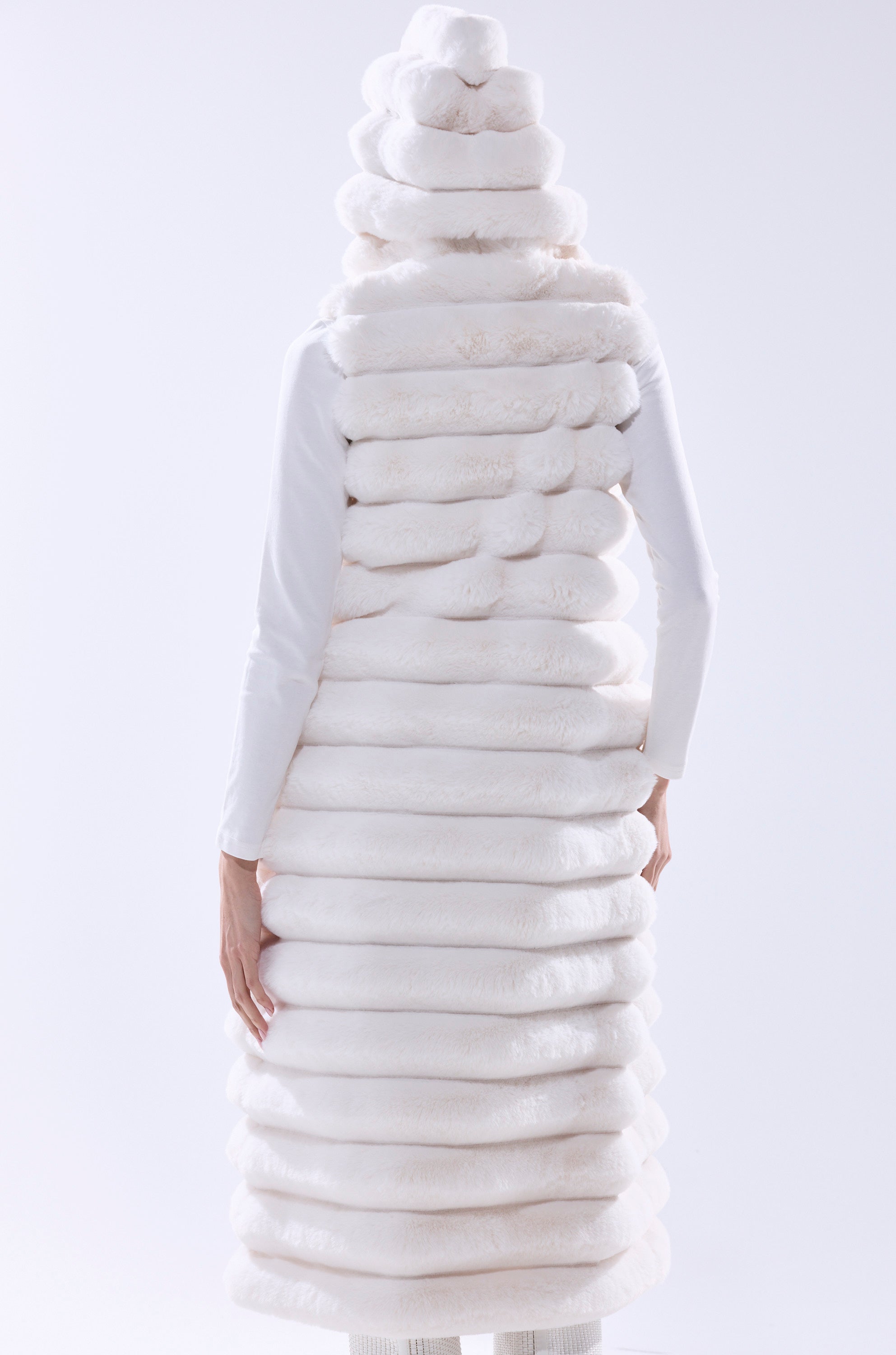 A person faces away, wearing the POPPY FAUX FUR HOODED VEST featuring a sculptural, layered look with a fluffy longline silhouette and segmented design, paired with matching tiered headwear against a plain light background.