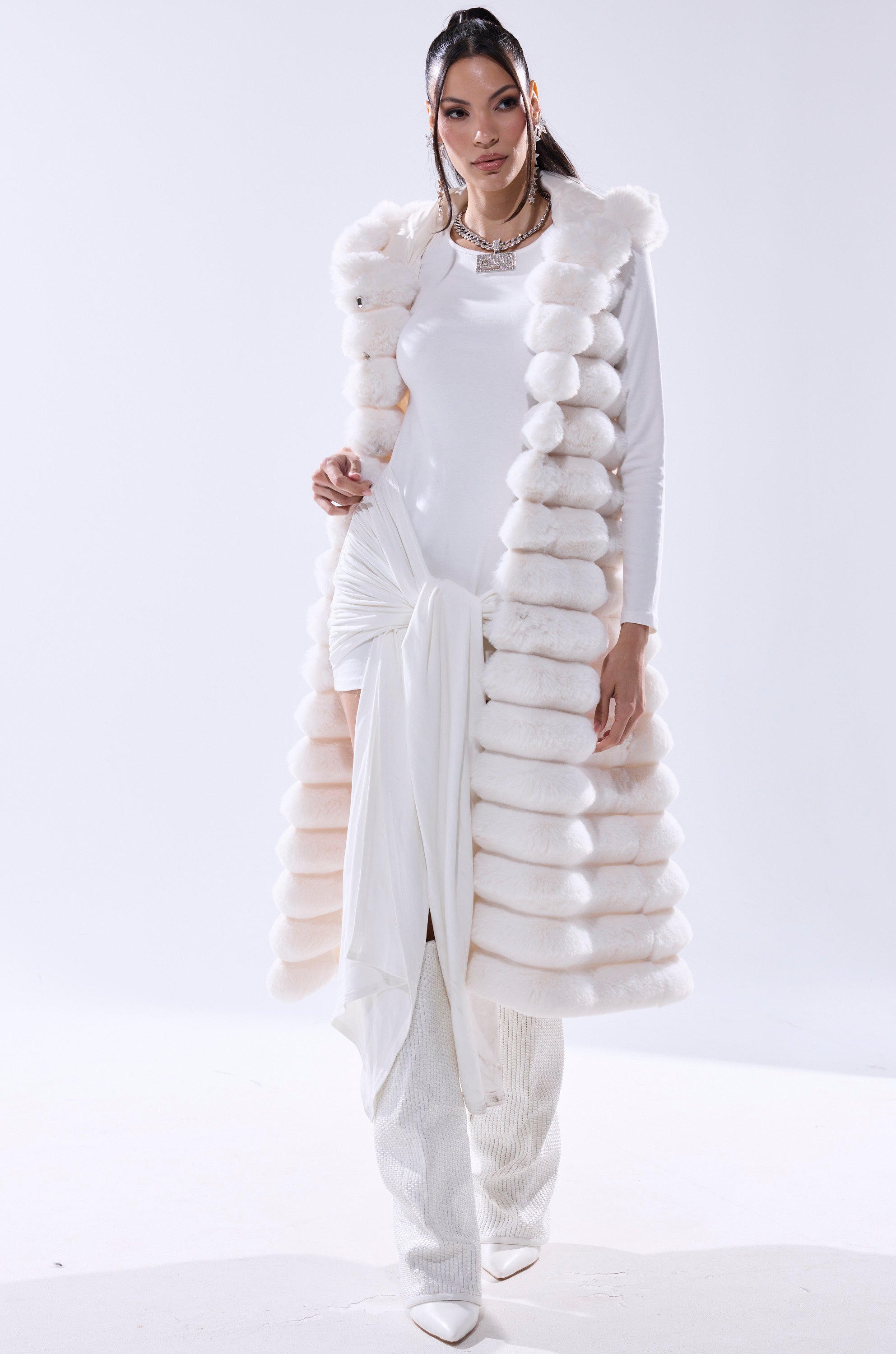 A woman poses in the POPPY FAUX FUR HOODED VEST, styled with wide-leg pants and a knotted accent. She finishes her chic winter look with high heels, jewelry, and slicked-back hair for a sophisticated ensemble.