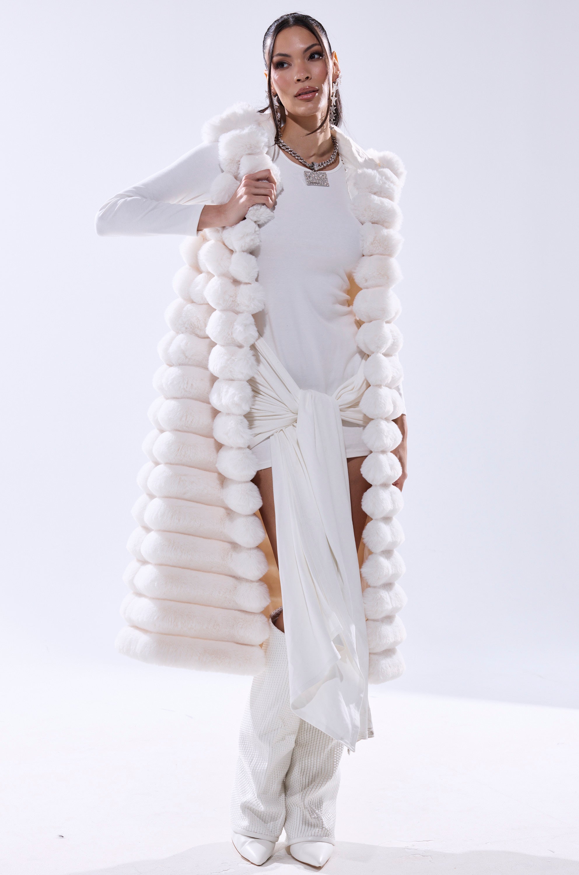 A woman exudes confidence in the POPPY FAUX FUR HOODED VEST, styled over a white draped dress with knee-high boots and layered silver necklaces, set against a light background.