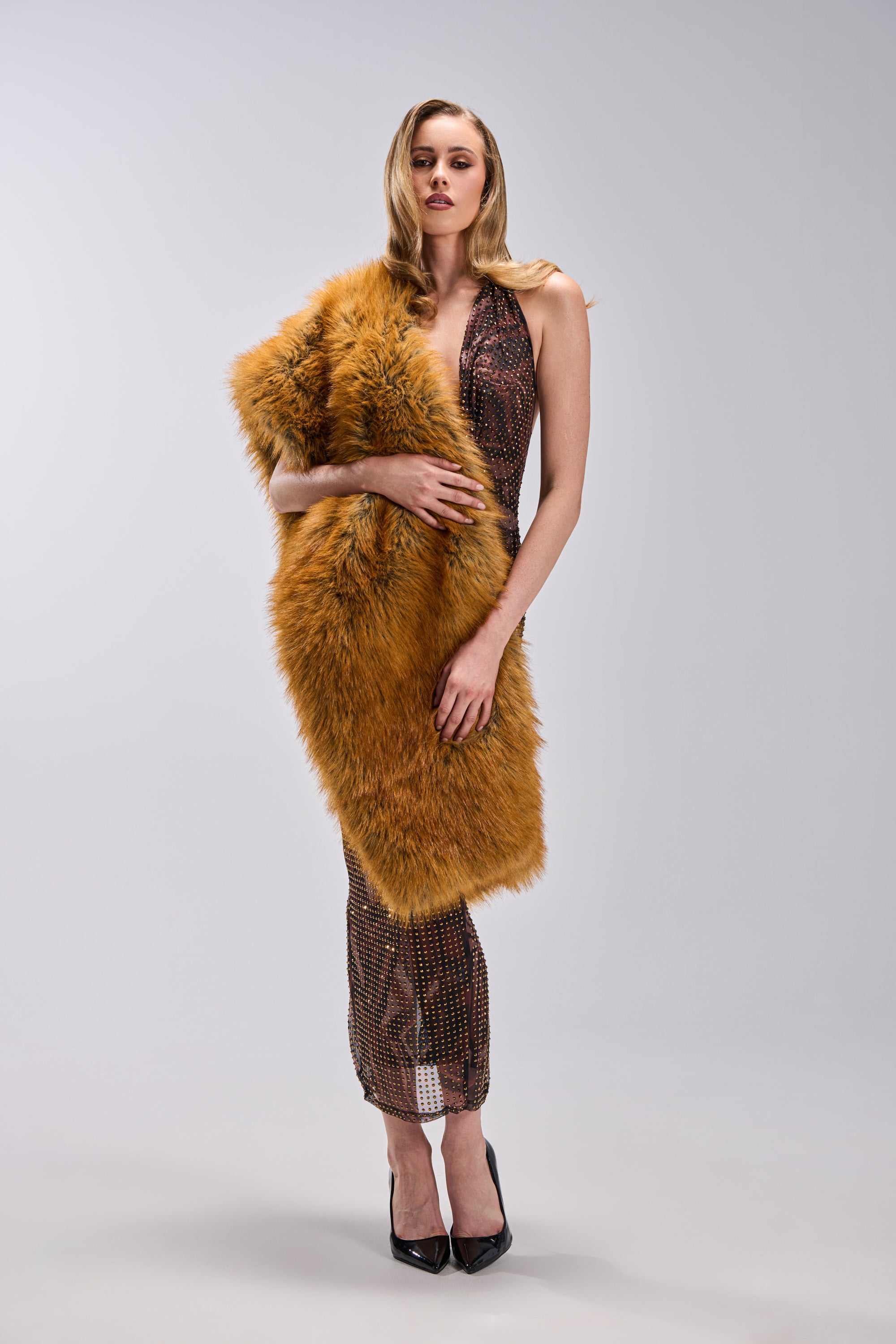A woman with long blonde hair and black heels stands against a plain background, wearing a long textured dress draped with the REALLY LIKE THAT FAUX FUR SCARF—a bold, fluffy orange piece that elevates her chic winter look.