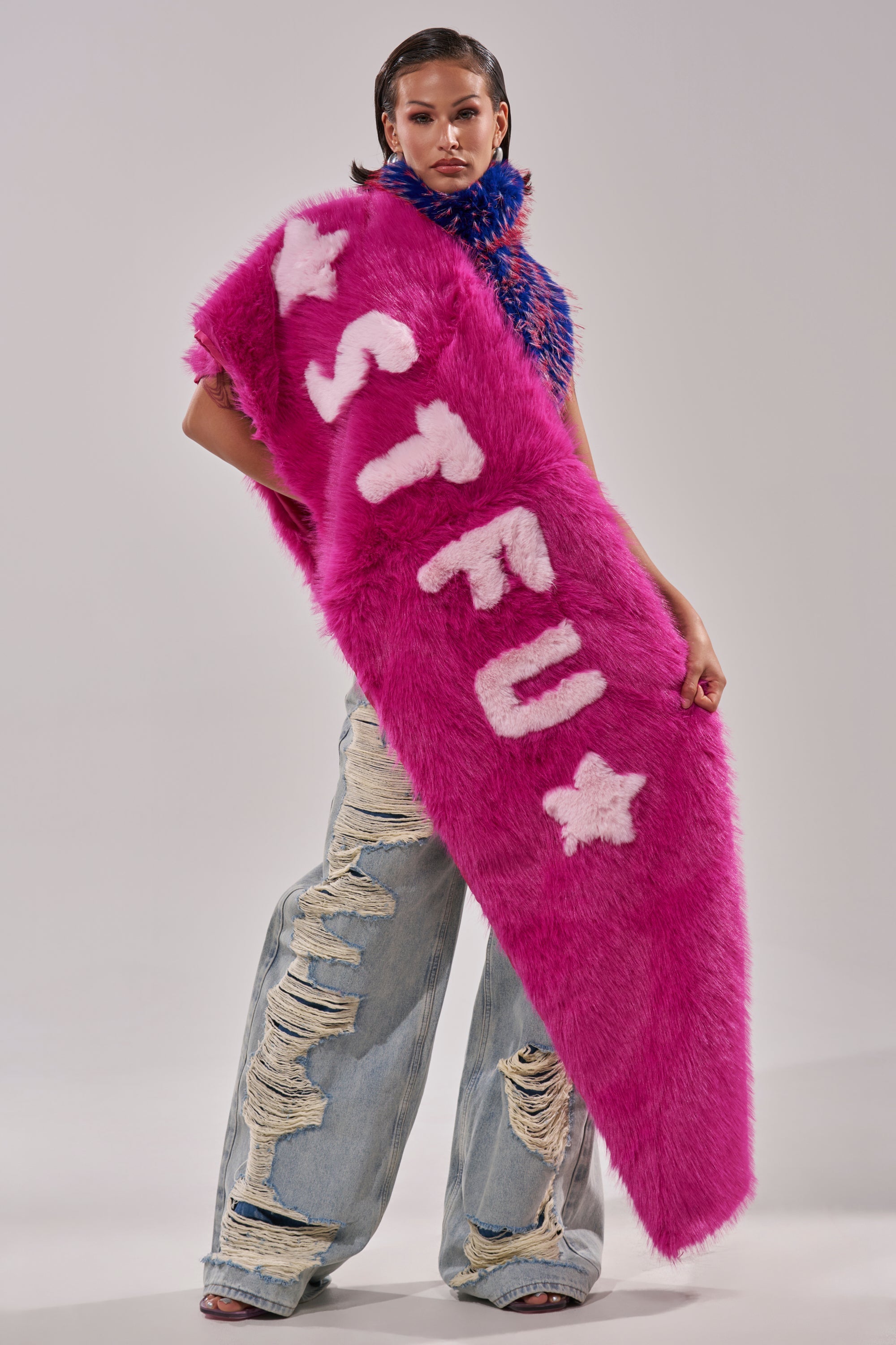 A person stands confidently in ripped jeans and the STFU FAUX FUR SCARF—an oversized fuchsia scarf with white "STFU" letters and stars—paired with a blue and pink textured top. 0